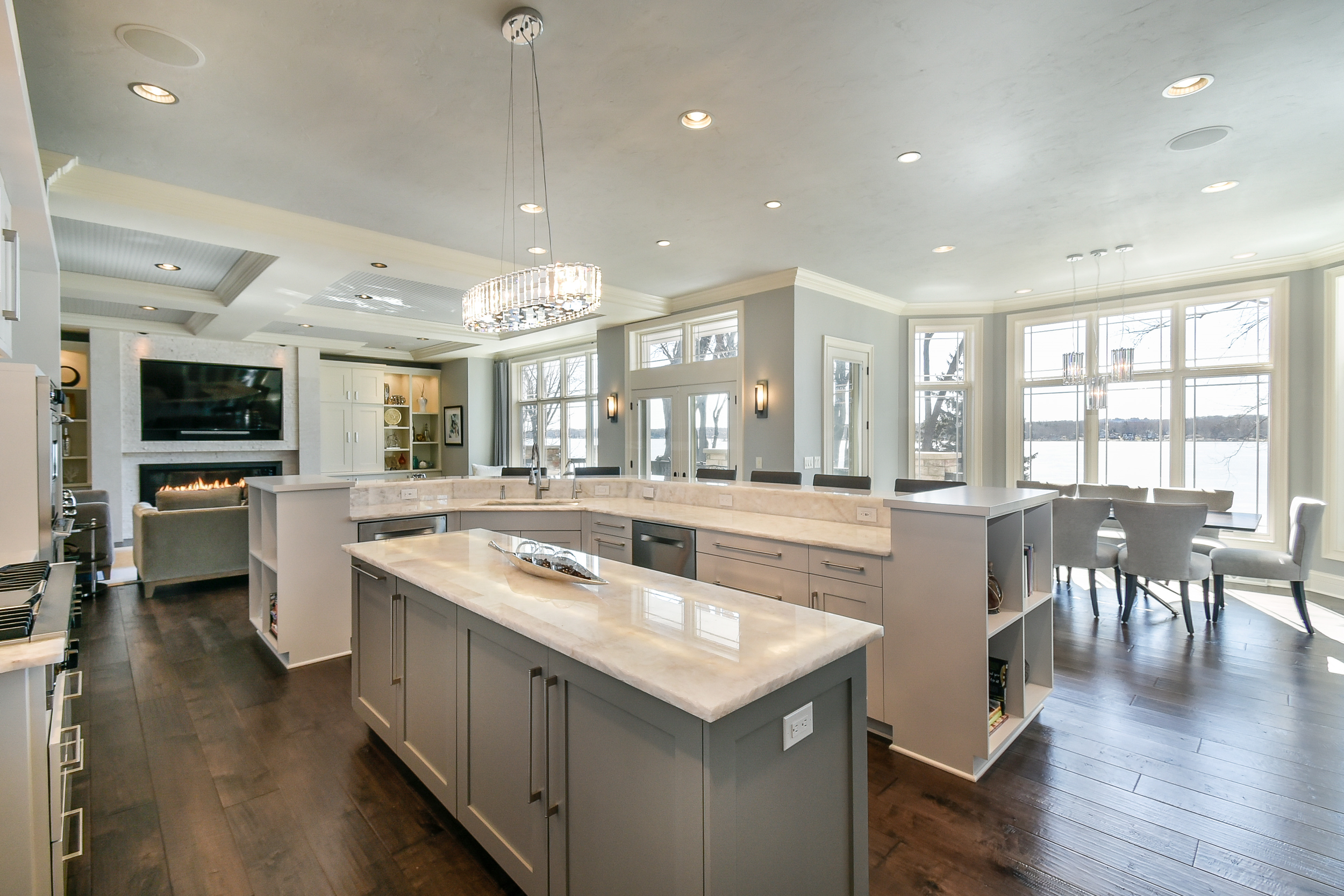 Bright, modern kitchen with gray cabinets, marble countertops, and a kitchen island. Large windows offer a scenic view of a body of water, with a dining area and a living room with a fireplace in the background.