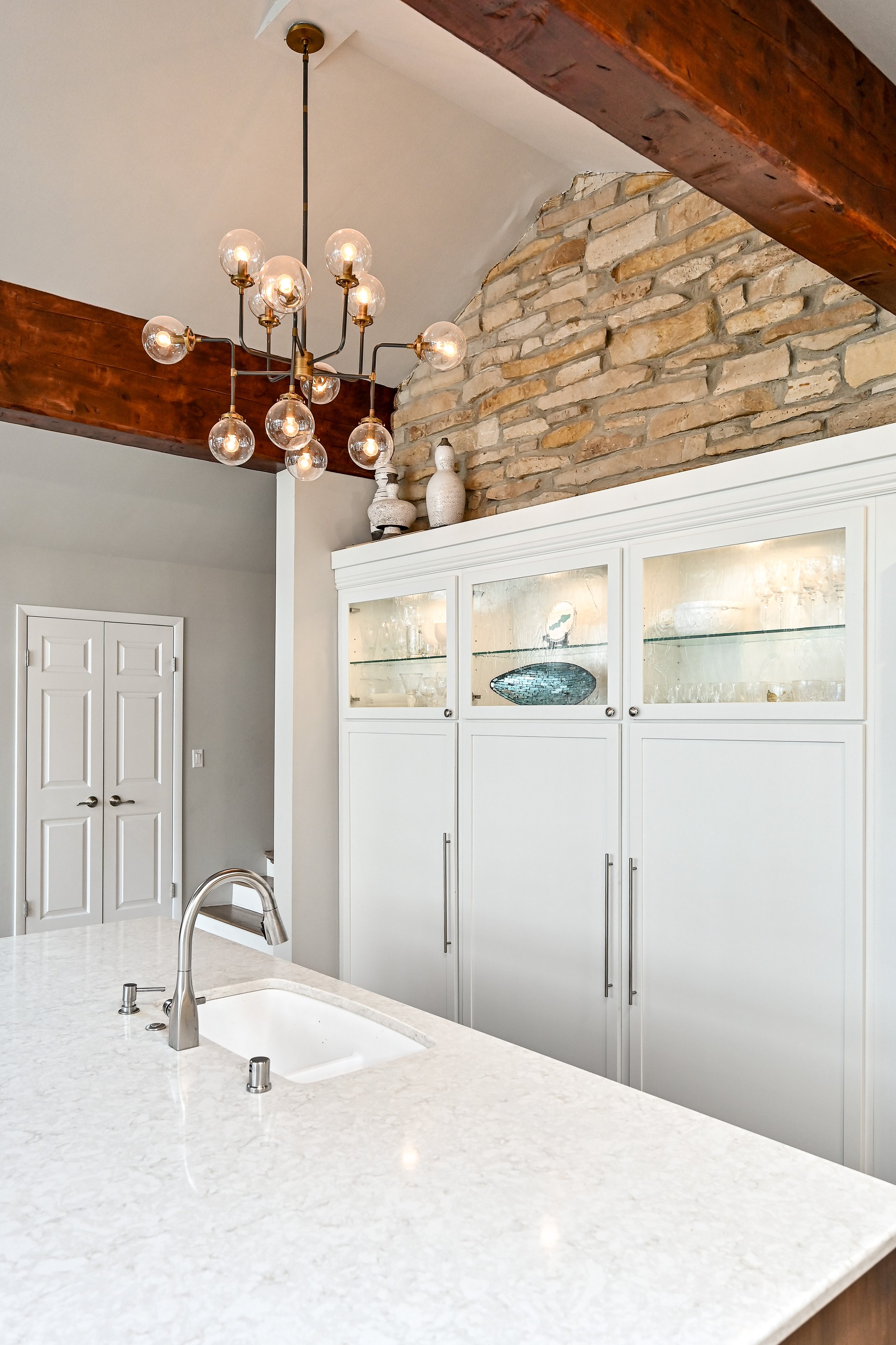 Modern kitchen with white cabinets, a marble countertop, a stainless steel faucet, and a brick chimney with wooden beams and a contemporary chandelier.