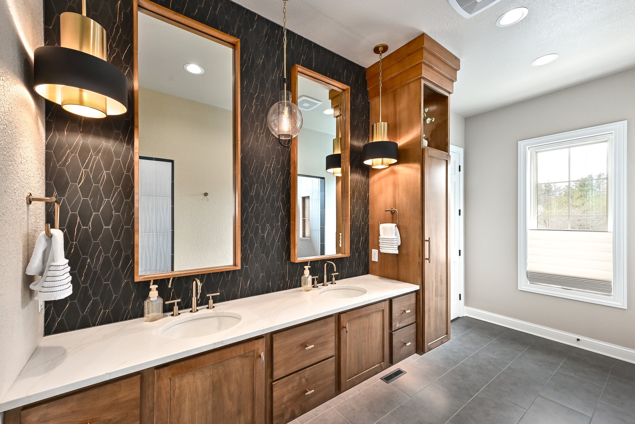 Bathroom with two sinks, marble countertop, wooden cabinets, black tiled wall with honeycomb pattern, two large mirrors, pendant lights, window with white frame, beige walls, gray tile floor.