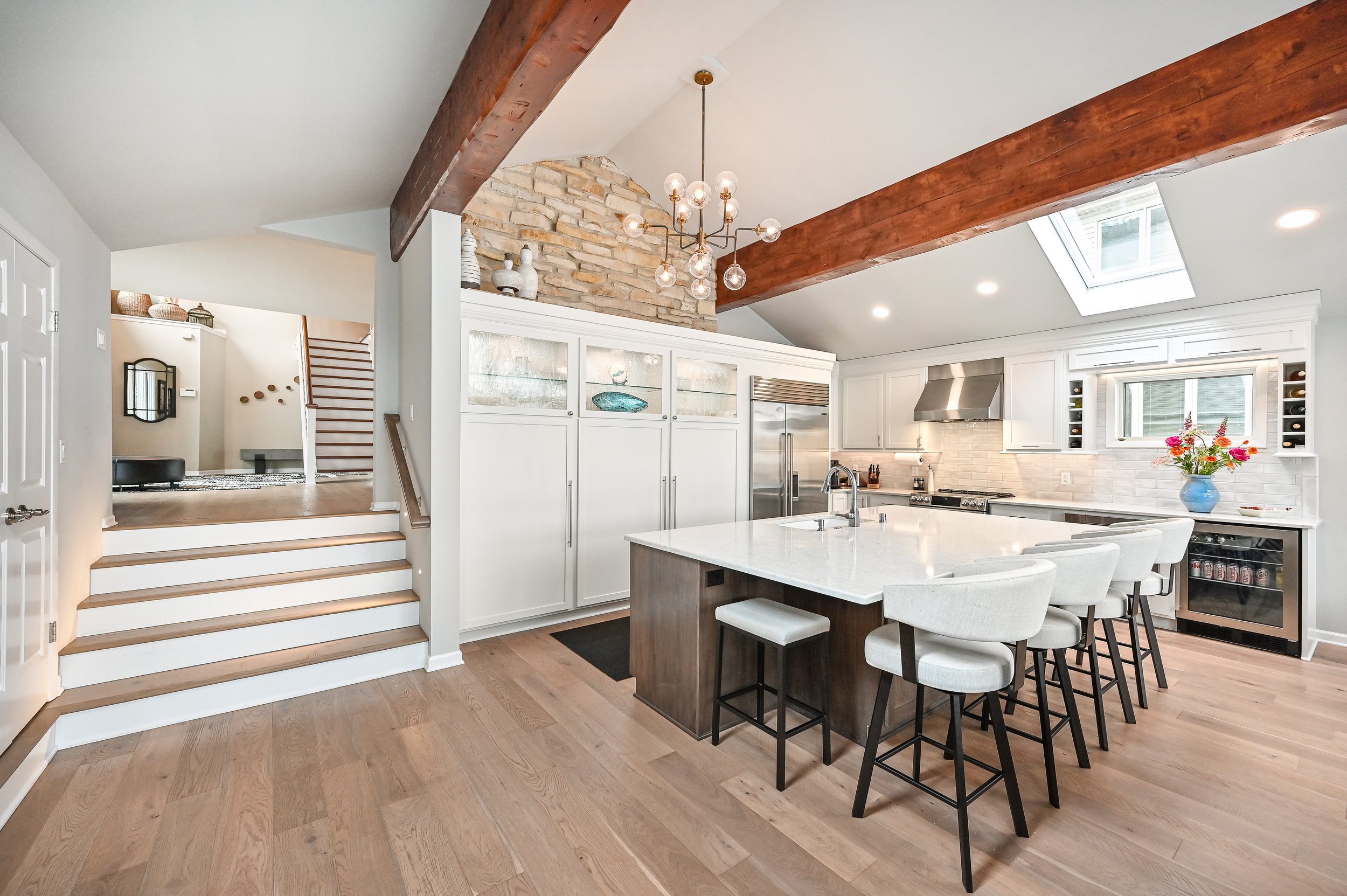 Modern kitchen with white cabinetry, a large island with a white countertop, stainless steel appliances, a skylight, a vase of flowers, and wooden beams on the ceiling.