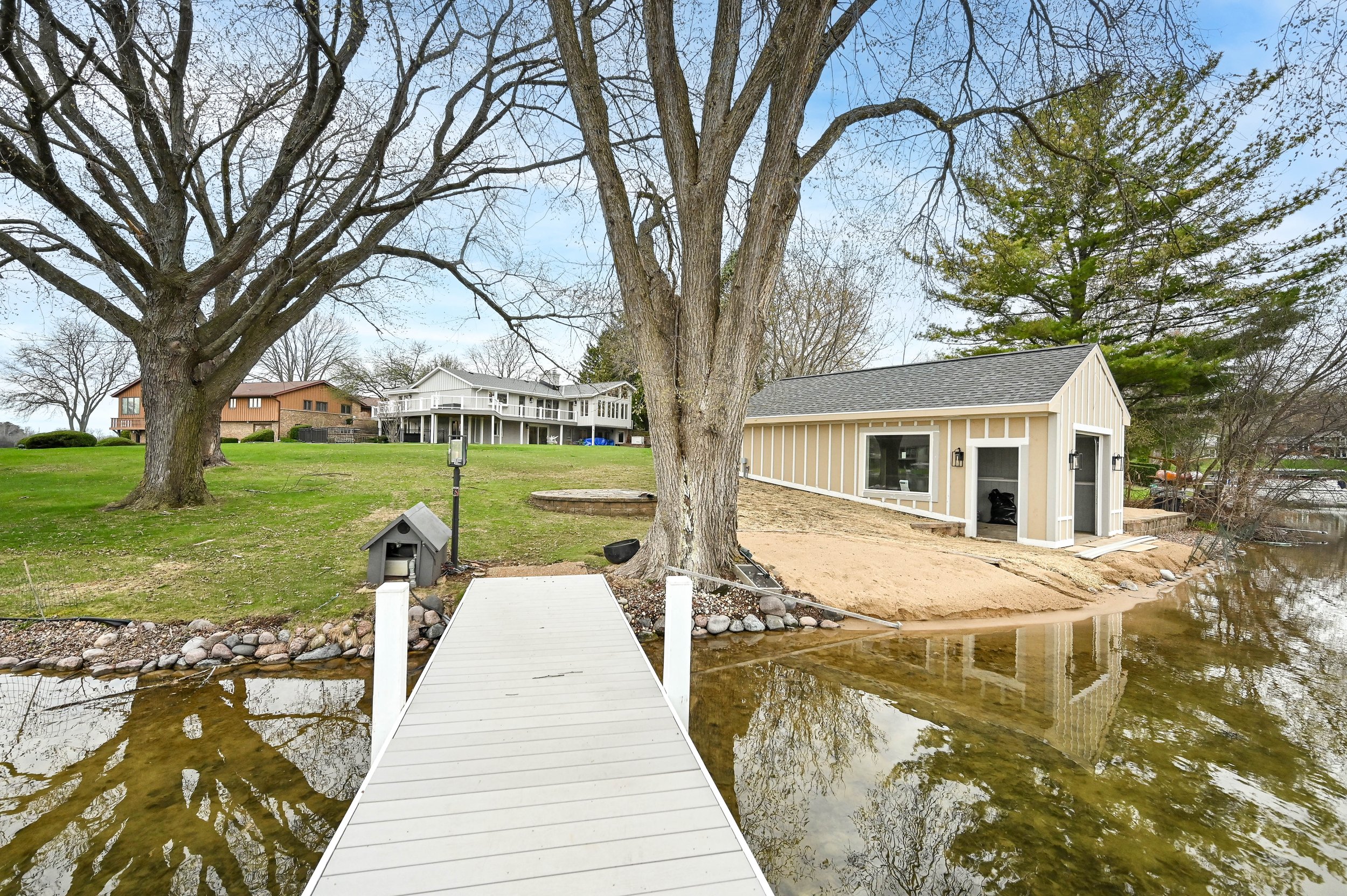 A backyard with a boat dock extending over a pond, grassy area with large trees, a small beige shed, and a multi-story house in the background on a partly cloudy day.