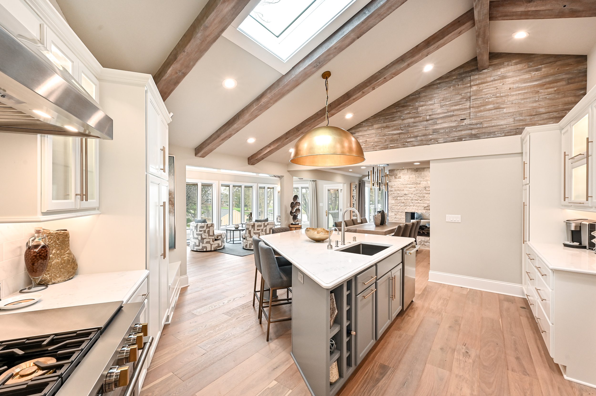 Open-concept kitchen with white cabinets, an island with gray cabinetry and a white countertop, wooden beams on the ceiling, a skylight, and a view into the living area with large windows and a sitting area.