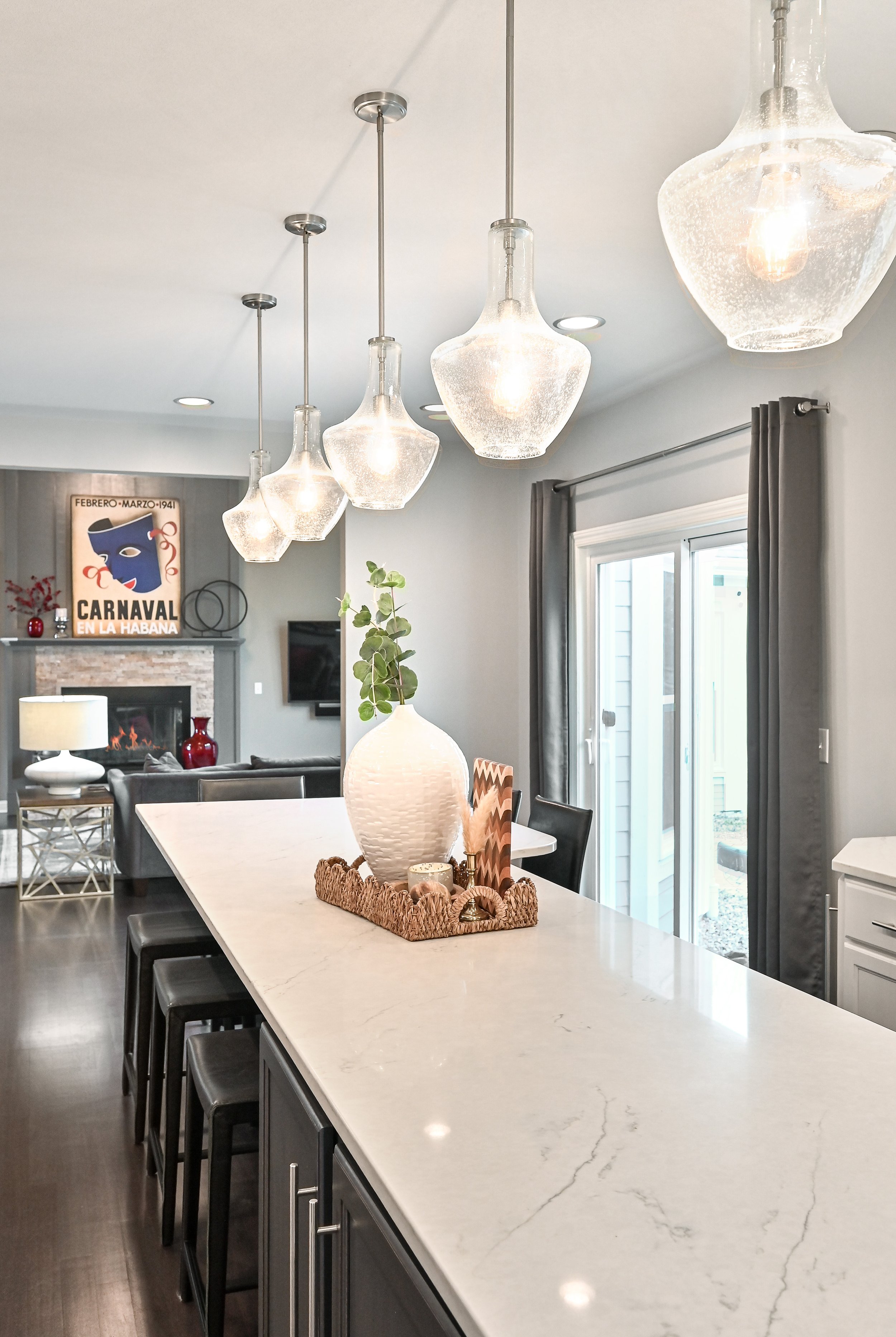 A modern kitchen and living room with a white marble countertop island, hanging glass pendant lights, a decorative tray with a vase and greenery, gray curtains, and a fireplace with a TV mounted above, in an open-plan living space.
