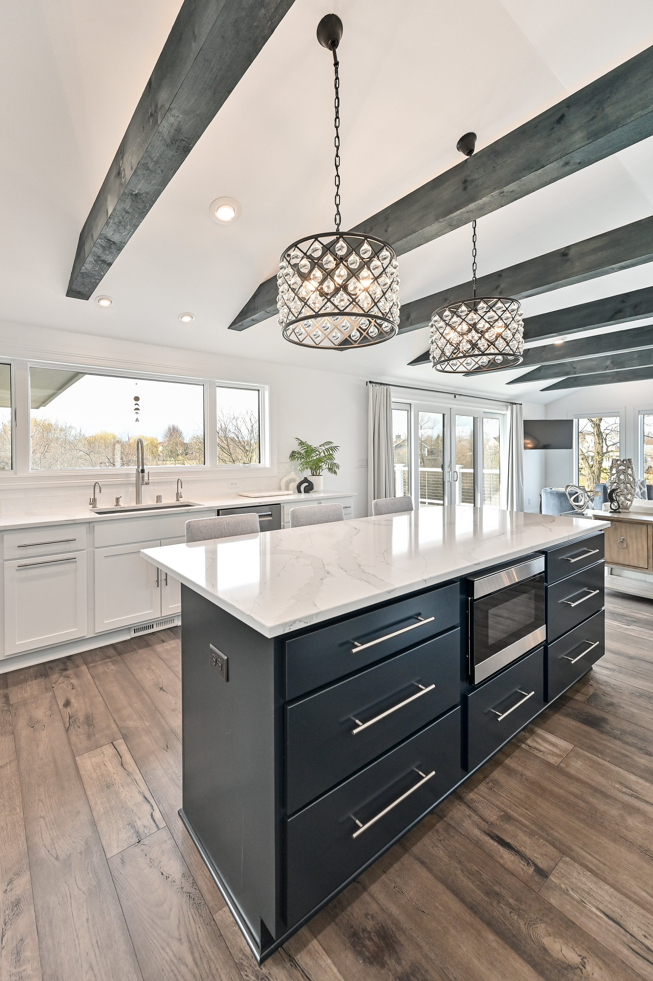Modern kitchen with a black island with drawers and a marble countertop, white kitchen cabinets, rustic wood flooring, large windows, and contemporary chandeliers with crystal accents hanging from a ceiling with black beams.