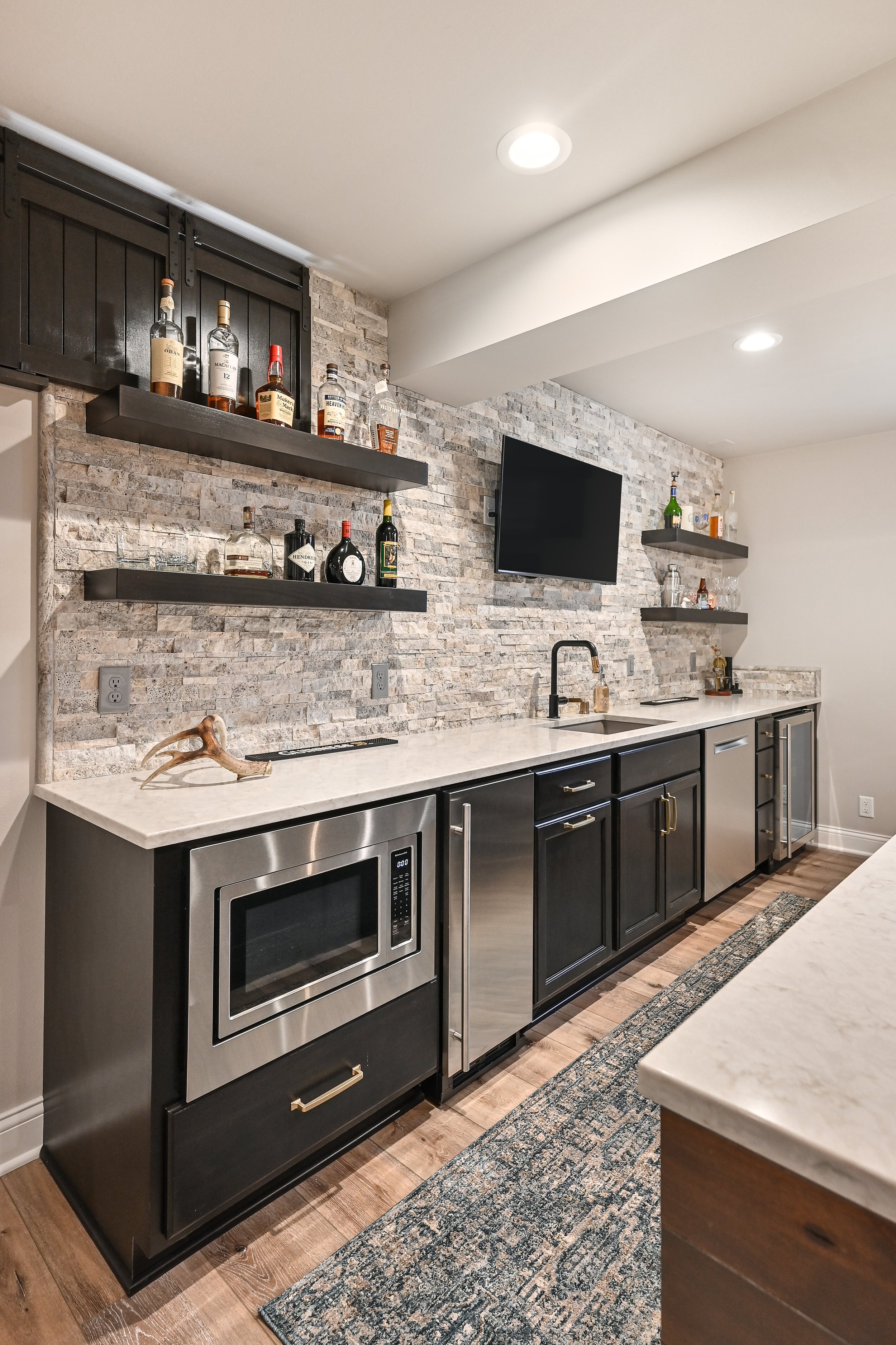 Modern kitchen with black cabinets, a marble countertop, a microwave, and open shelves with liquor bottles and glassware.