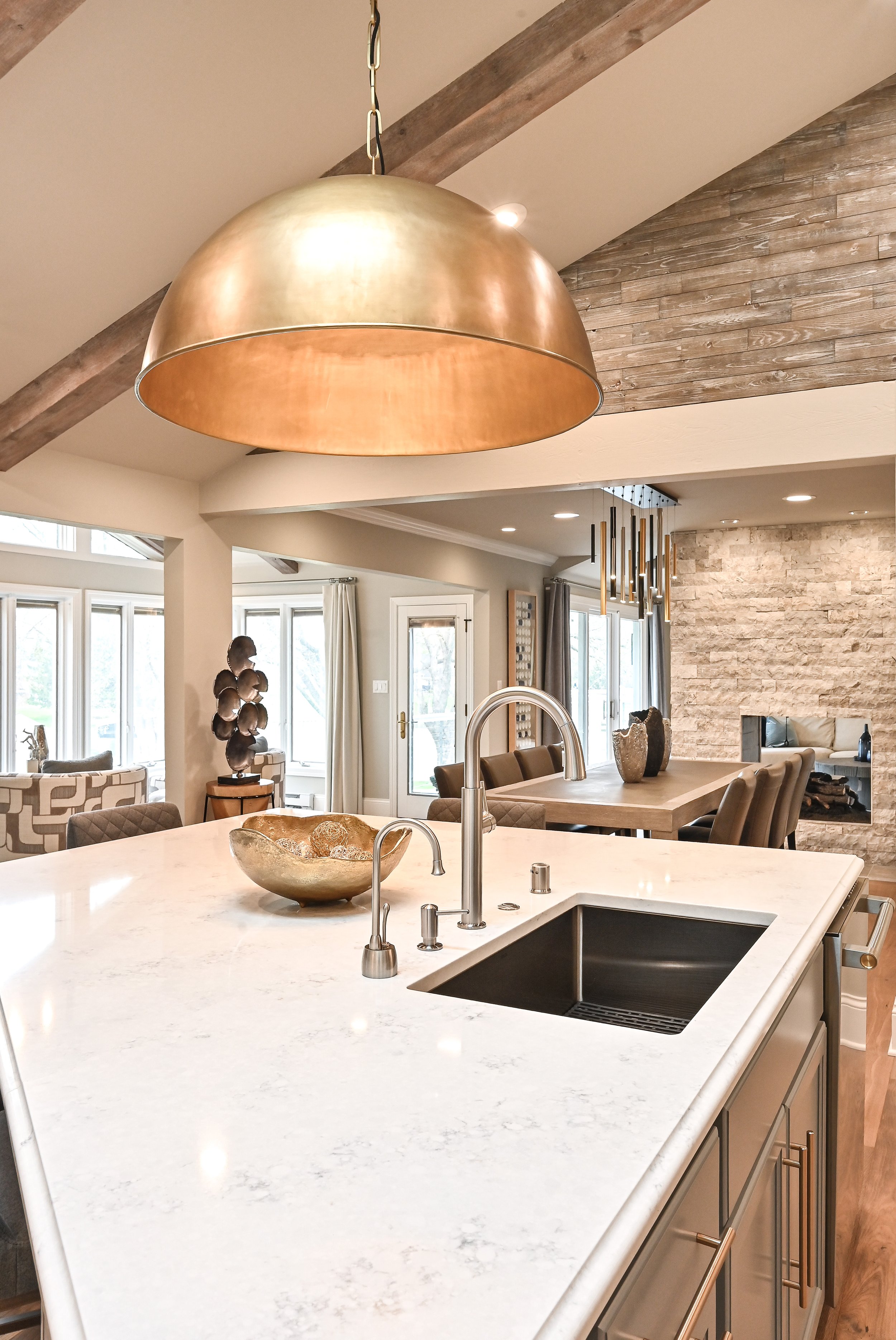 Modern kitchen with white marble countertop, gold pendant lighting, and open living and dining area with large windows and neutral decor.