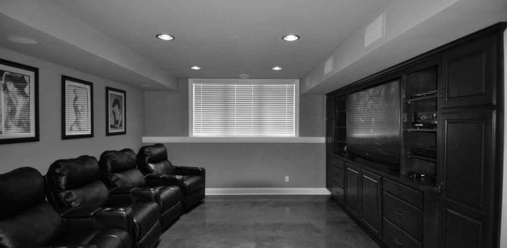 Basement living room with four black leather recliners, framed black and white photos on the wall, and a large built-in entertainment center with a TV, shelves, and drawers, in black finish.