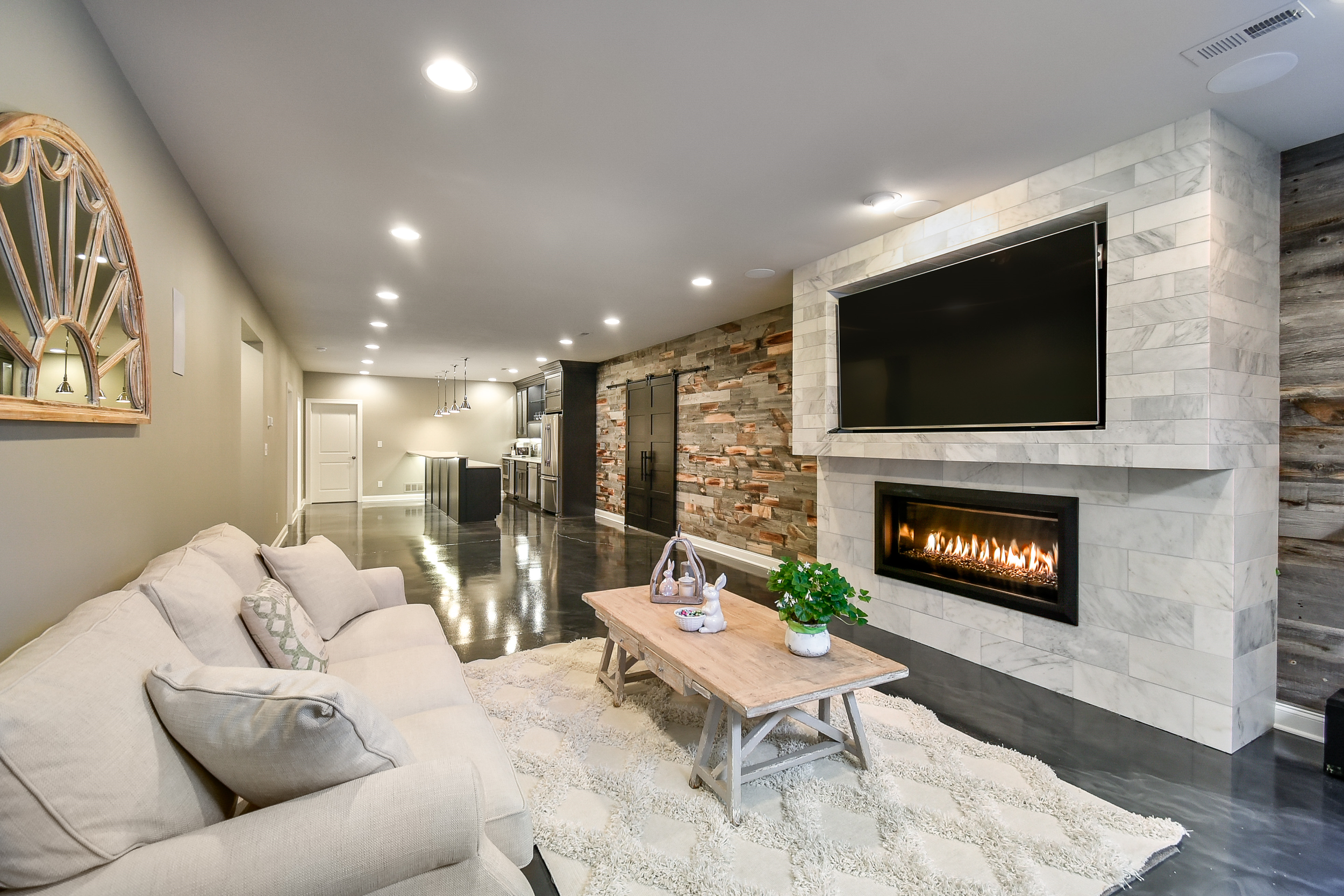 Living room with a beige sofa, a wooden coffee table with decorative items and a plant, a wall-mounted TV above a fireplace, a wood accent wall, and an open kitchen in the background.