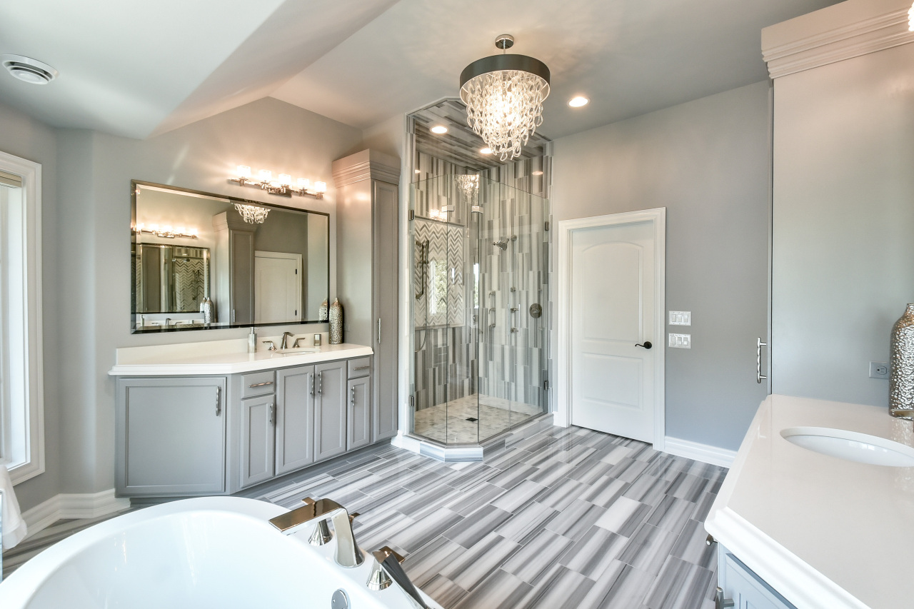 Modern bathroom with gray cabinets, a large mirror, a glass-enclosed shower with gray marble tiles, a chandelier, a bathtub, and striped gray and white floor tiles.