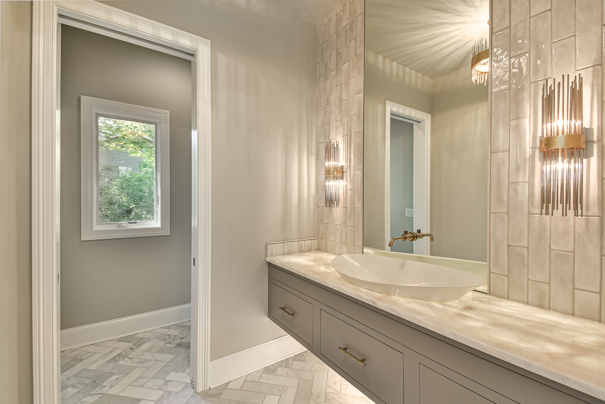 Modern bathroom with a large mirror, a vessel sink, and elegant sconces on the wall. A window shows greenery outside.