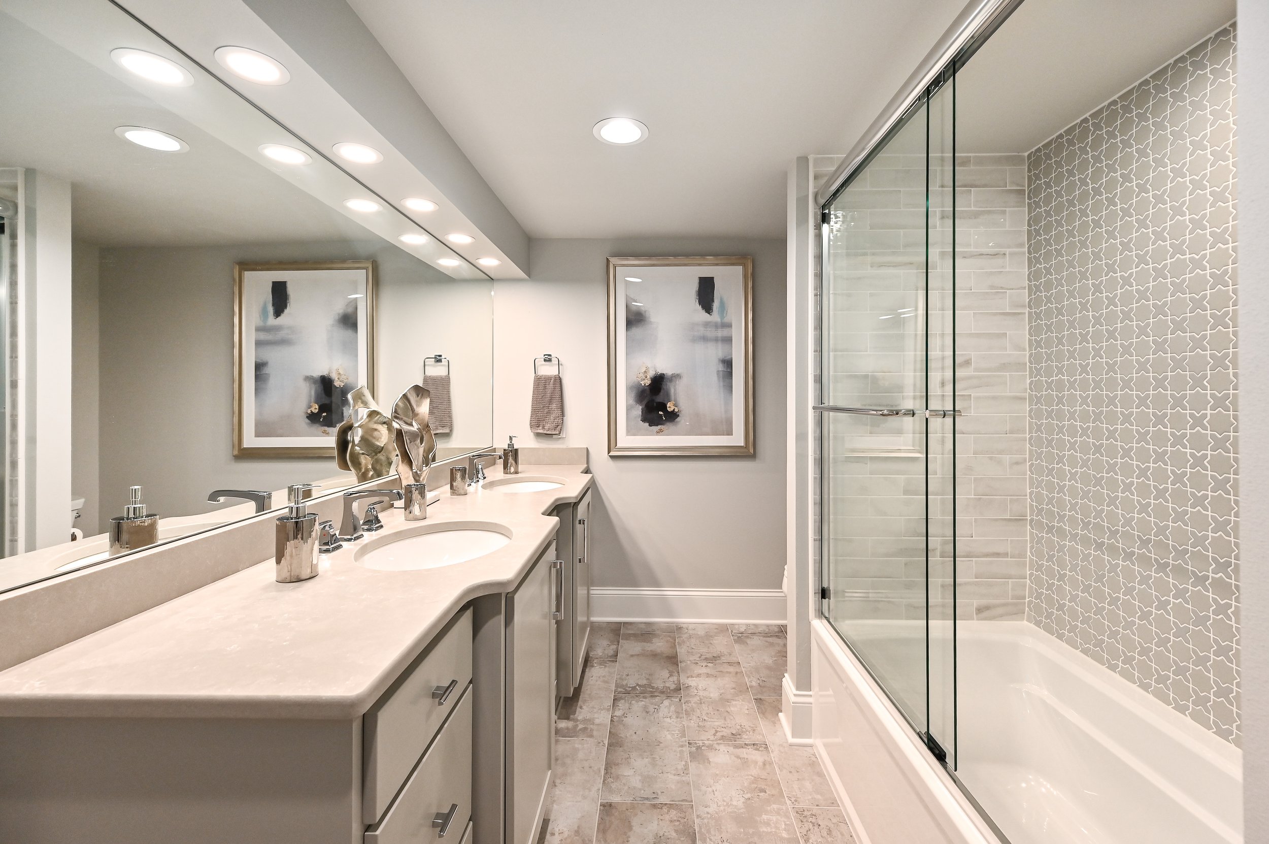 Modern bathroom with a double vanity sink, large mirror, abstract artwork, towels, and a glass-enclosed shower with decorative wall tiles.