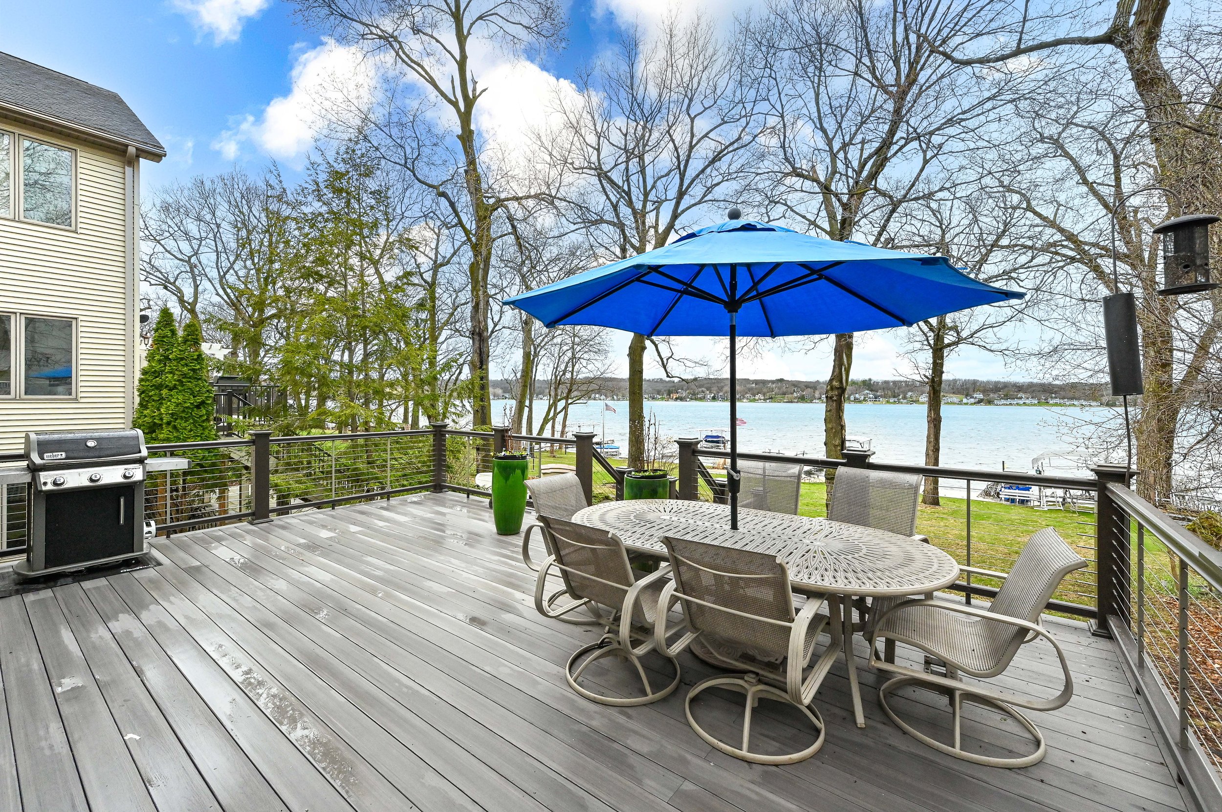 Deck overlooking a lake with outdoor furniture, a blue umbrella, trees, and docked boats in the distance.