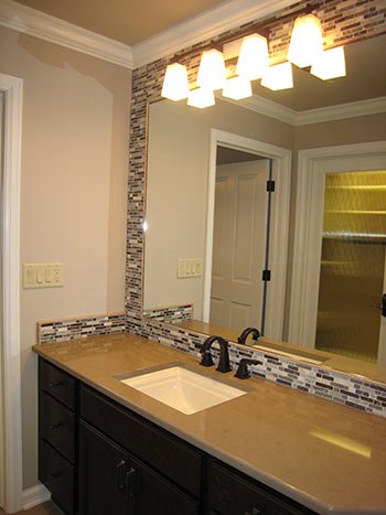 Bathroom vanity with a beige countertop, black cabinet, and a rectangular sink. Above the mirror are four wall-mounted lights, and to the right is a door with a glass panel.