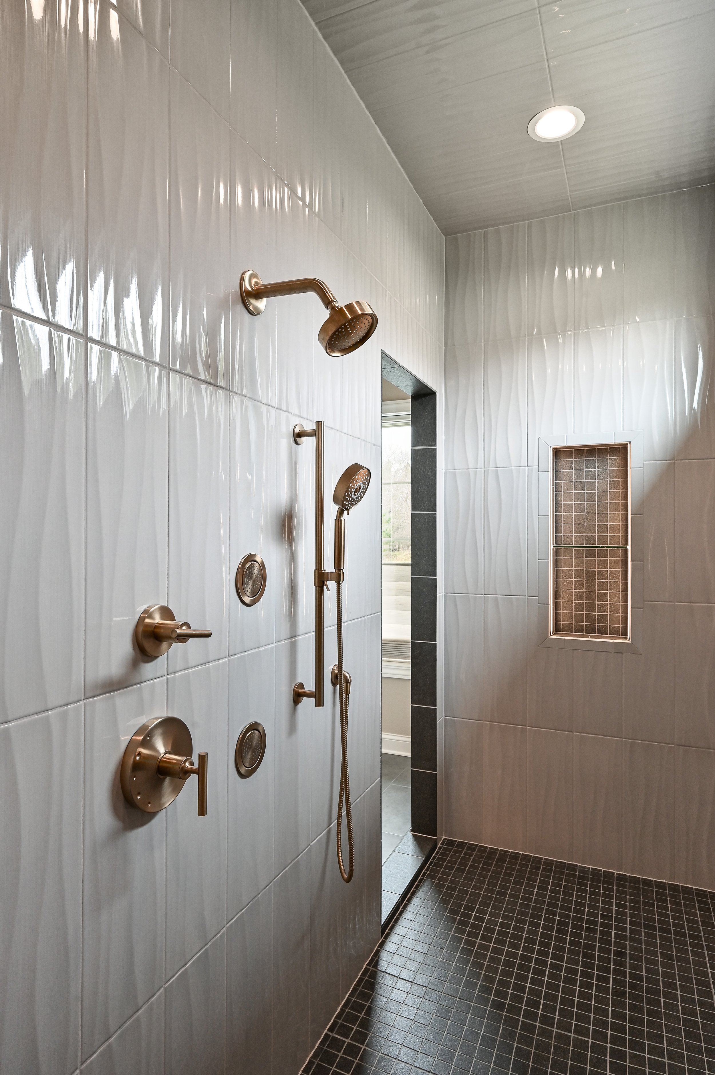 A modern shower with multiple showerheads and controls, tiled walls, a small window, and a ceiling light.
