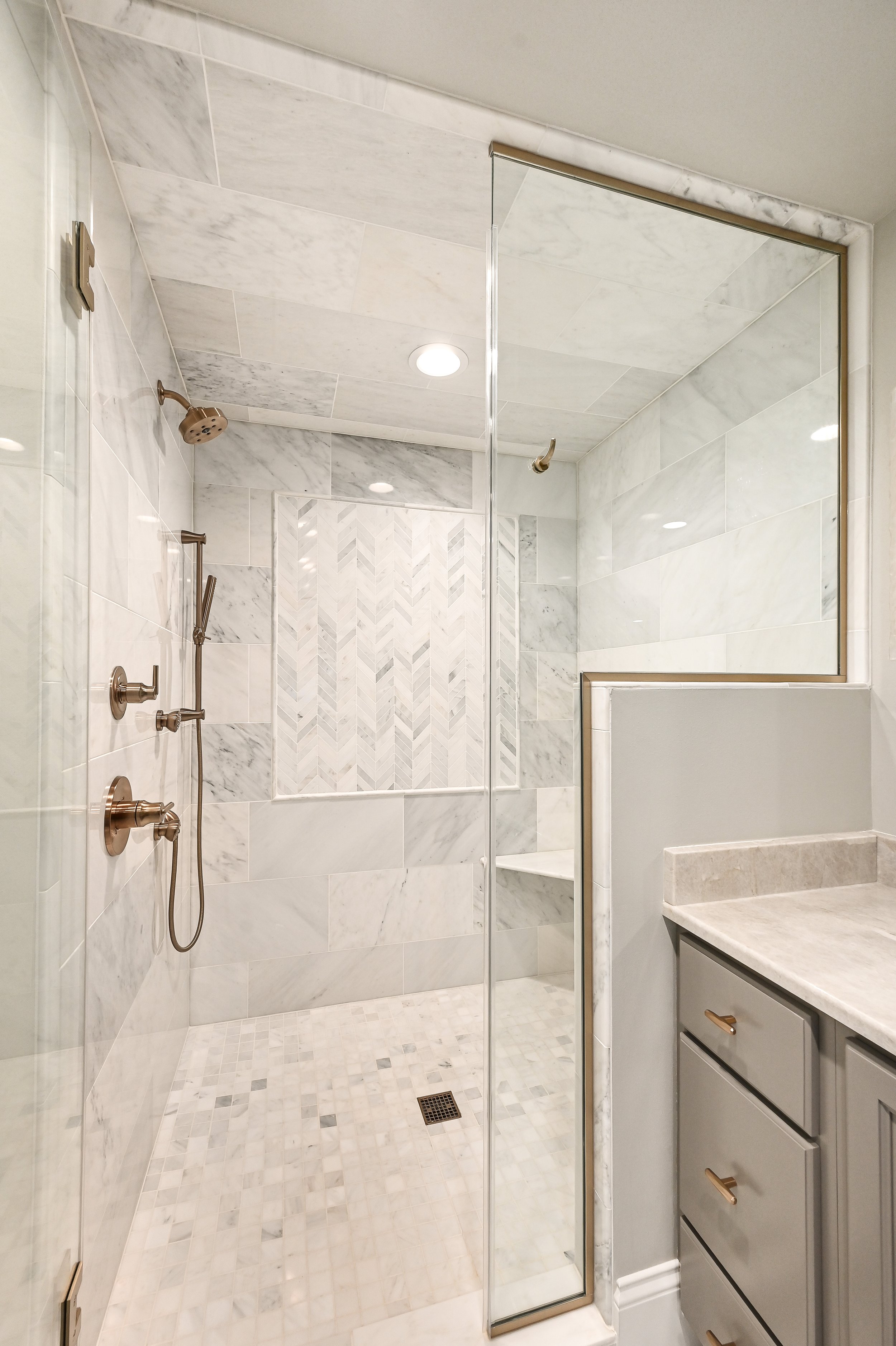 Modern walk-in shower with marble tiles, a dual shower head, and a built-in shelf, next to a bathroom vanity with a marble countertop and gray cabinets.