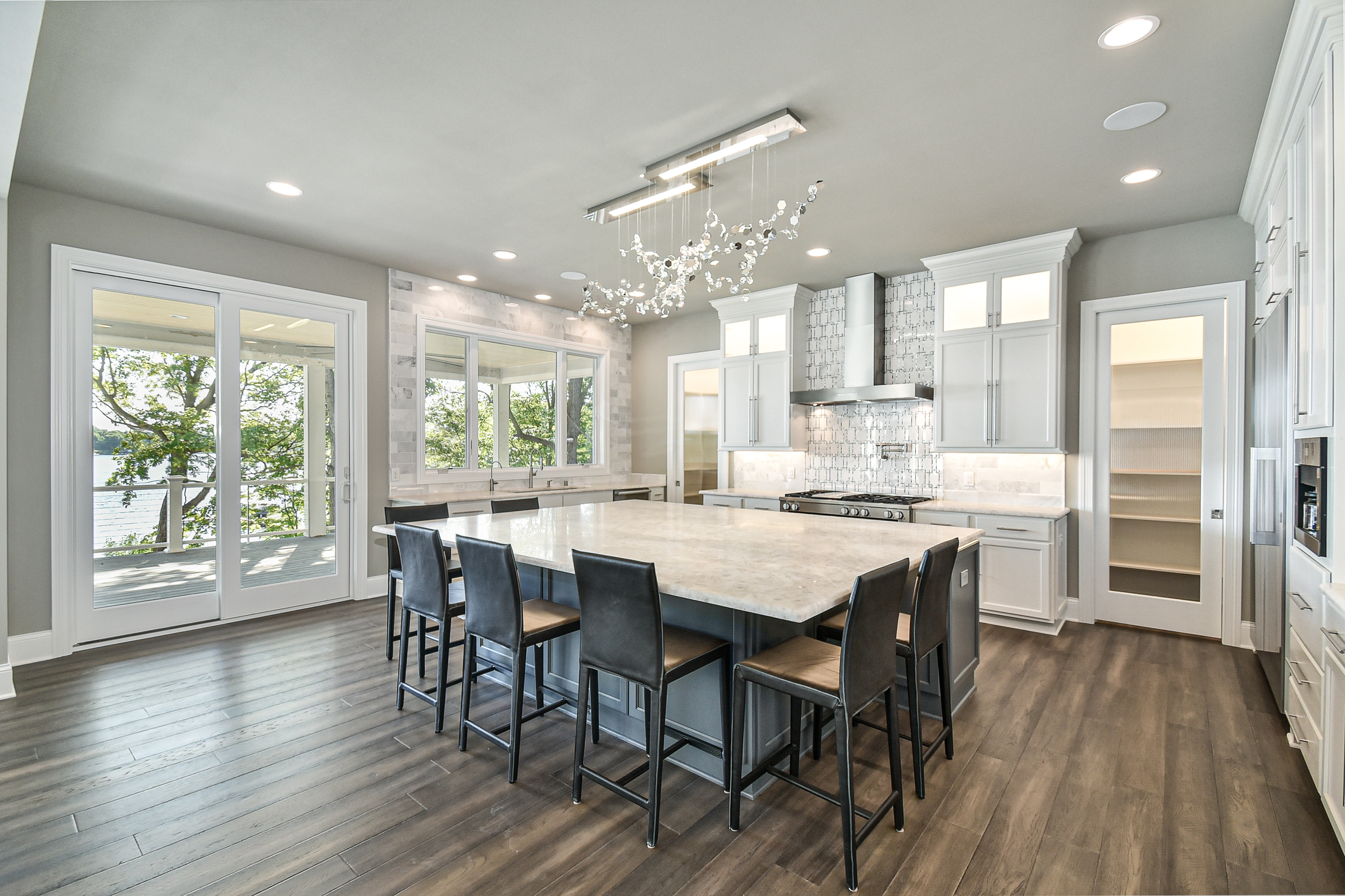 Modern kitchen with large island, white cabinets, stainless steel appliances, sliding glass door, and windows overlooking a lake, with a wood floor and decorative chandelier.