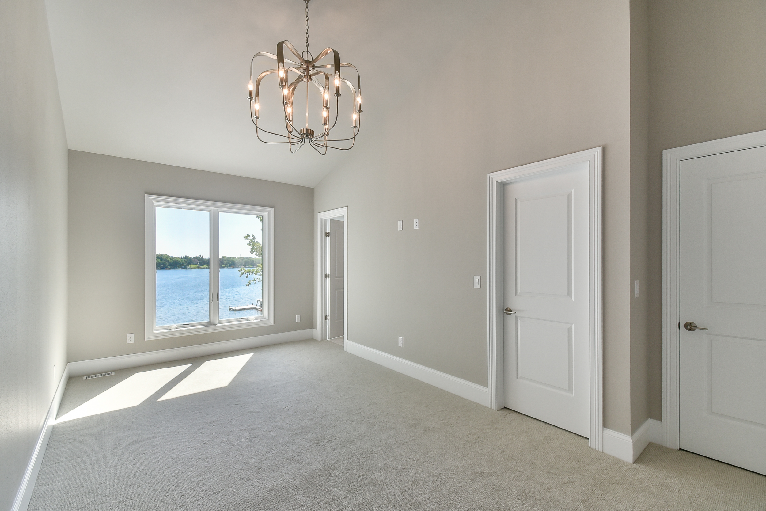 Empty room with large window overlooking a body of water, white walls, neutral carpet, chandelier hanging from ceiling, multiple white doors, bright natural light.