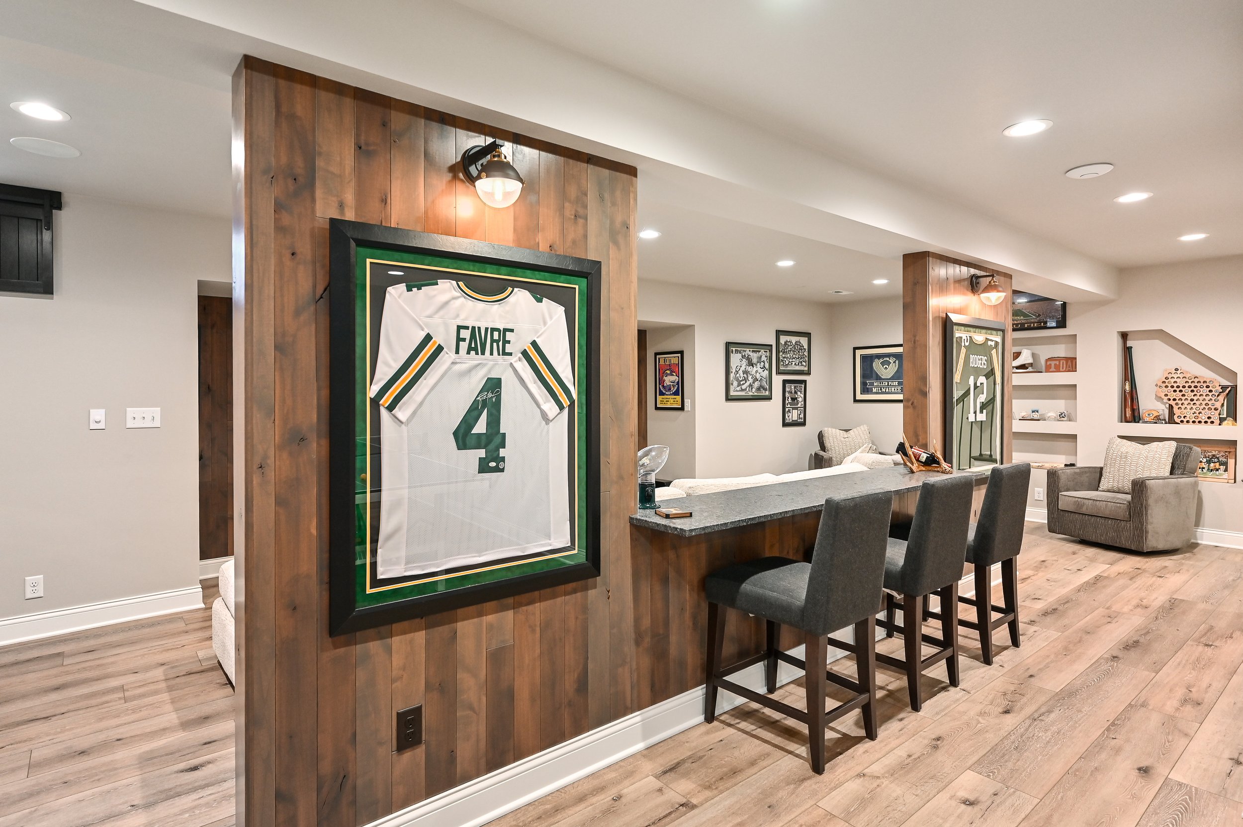 A basement living space decorated with framed sports jerseys and memorabilia, featuring wooden accent walls and hardwood floors.