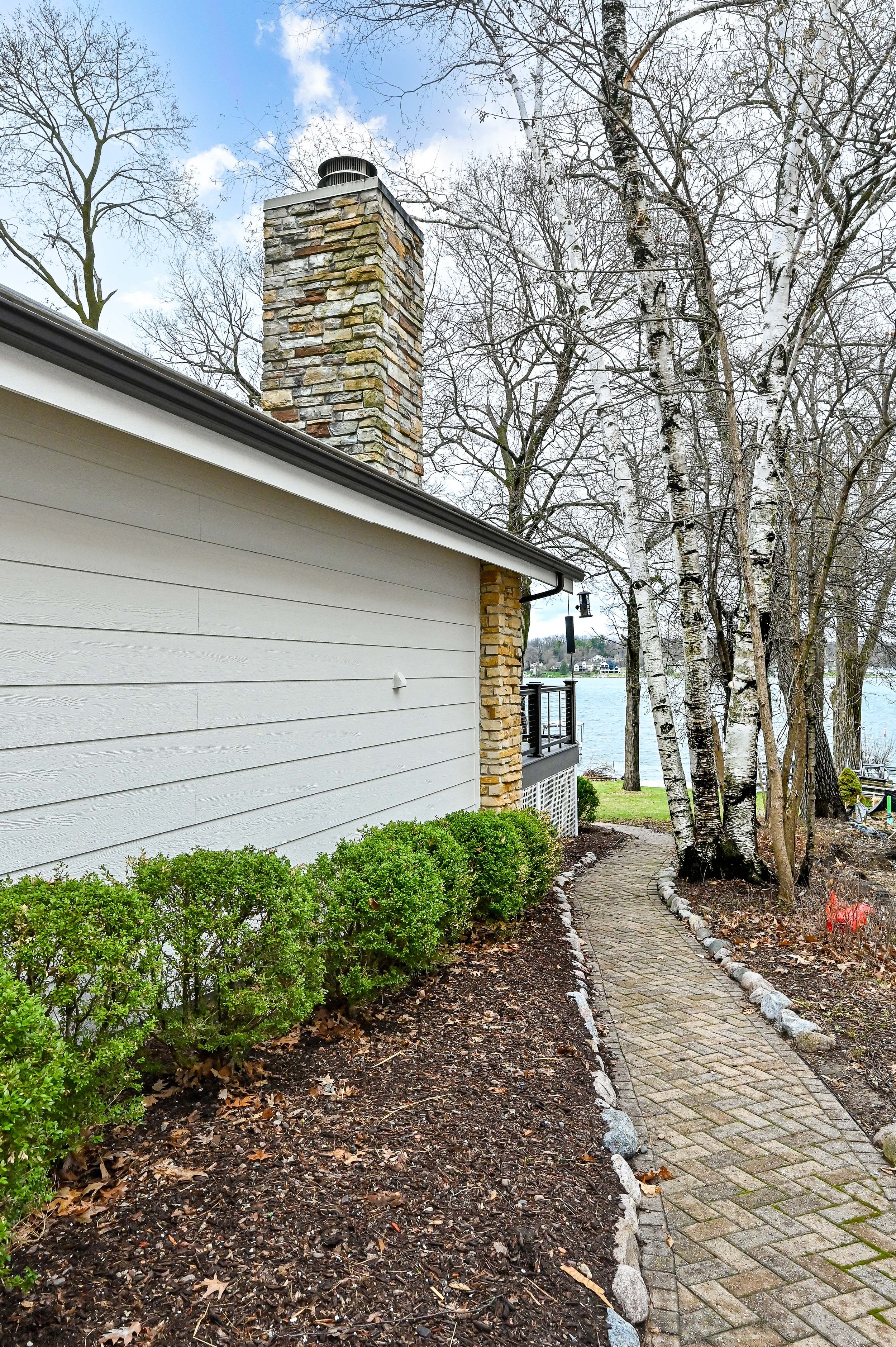 A house with white siding and a stone chimney next to a paved walkway, with trees and a lake in the background.
