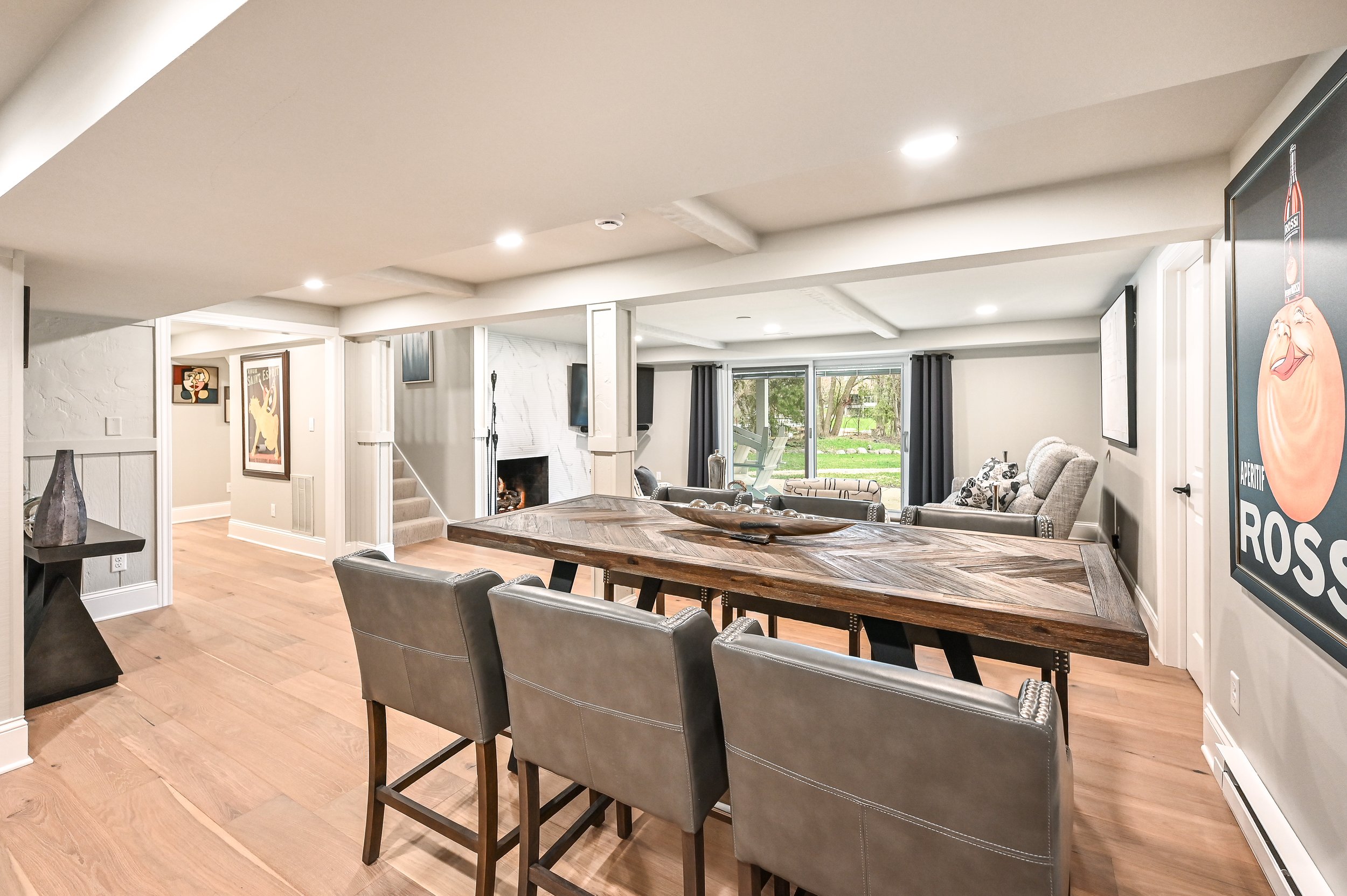 Spacious basement living area with a wooden dining table, leather chairs, and a sitting area with armchairs near a sliding glass door leading outside.