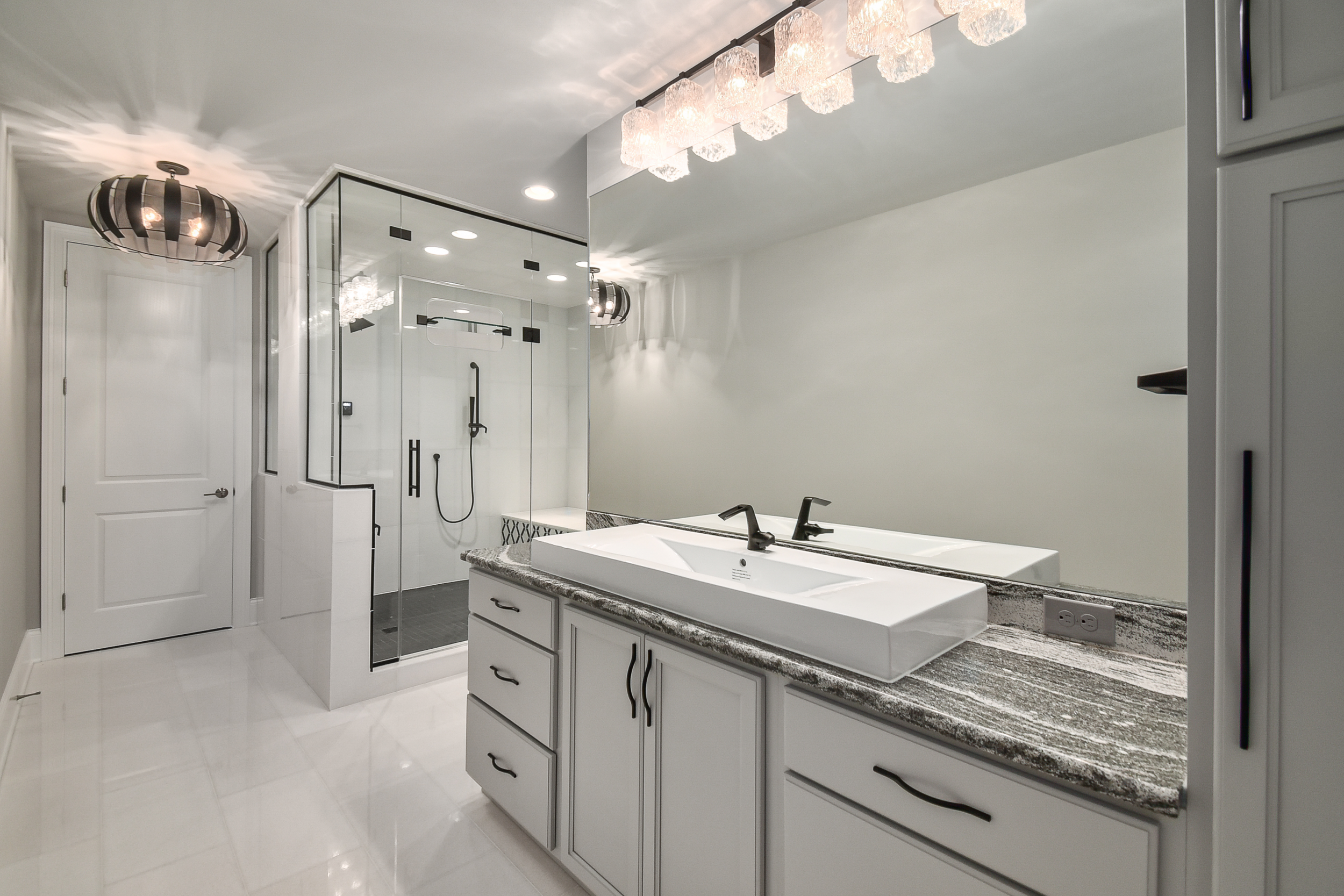 Modern bathroom with a white vanity, vessel sink, large mirror, glass shower with black fixtures, decorative lighting fixtures, and white tiled floor and walls.