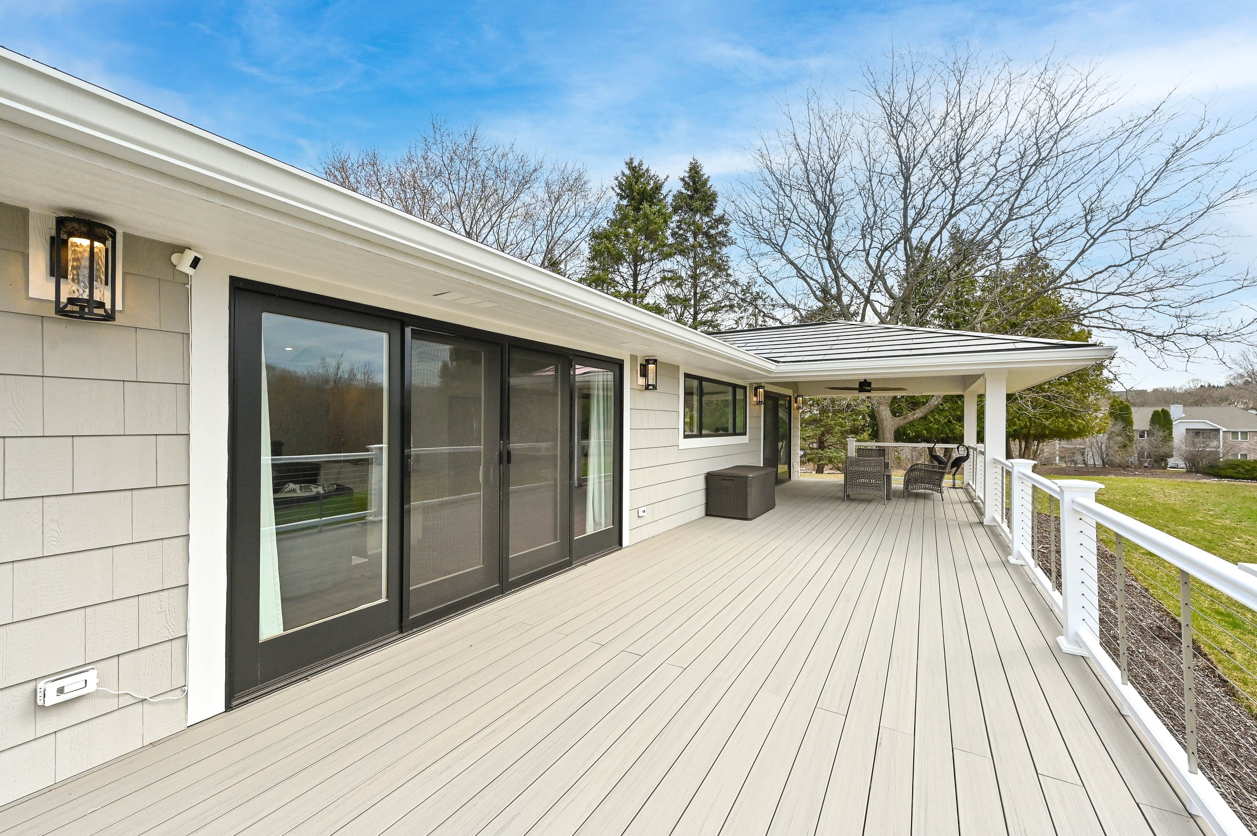 Large outdoor deck attached to a house, featuring sliding glass doors, outdoor furniture, ceiling fan, and wall-mounted lights, with a view of trees and neighboring houses.