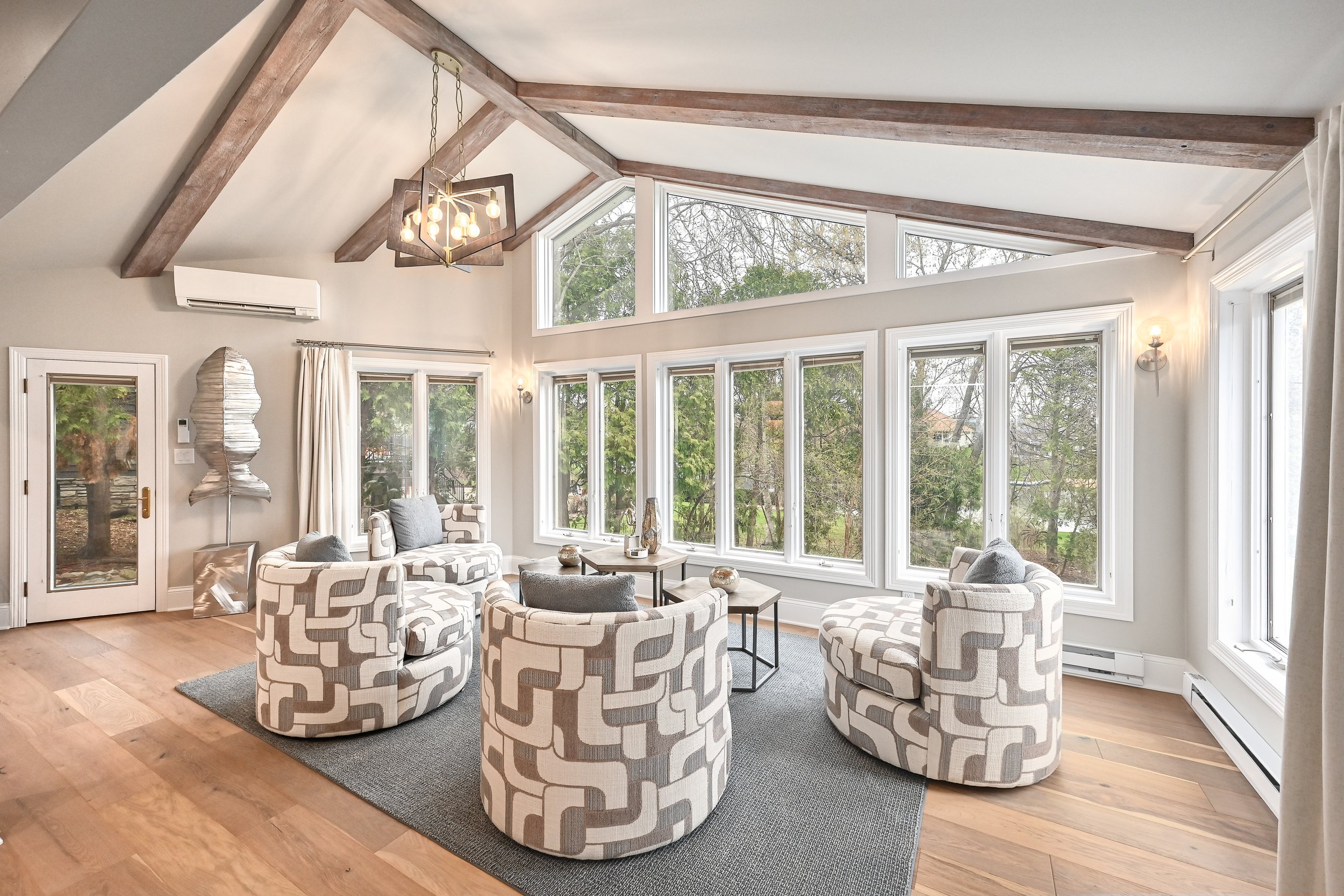Bright living room with large bay windows, four patterned armchairs arranged in a circle, wooden floors, and a modern chandelier hanging from a vaulted ceiling with wooden beams.