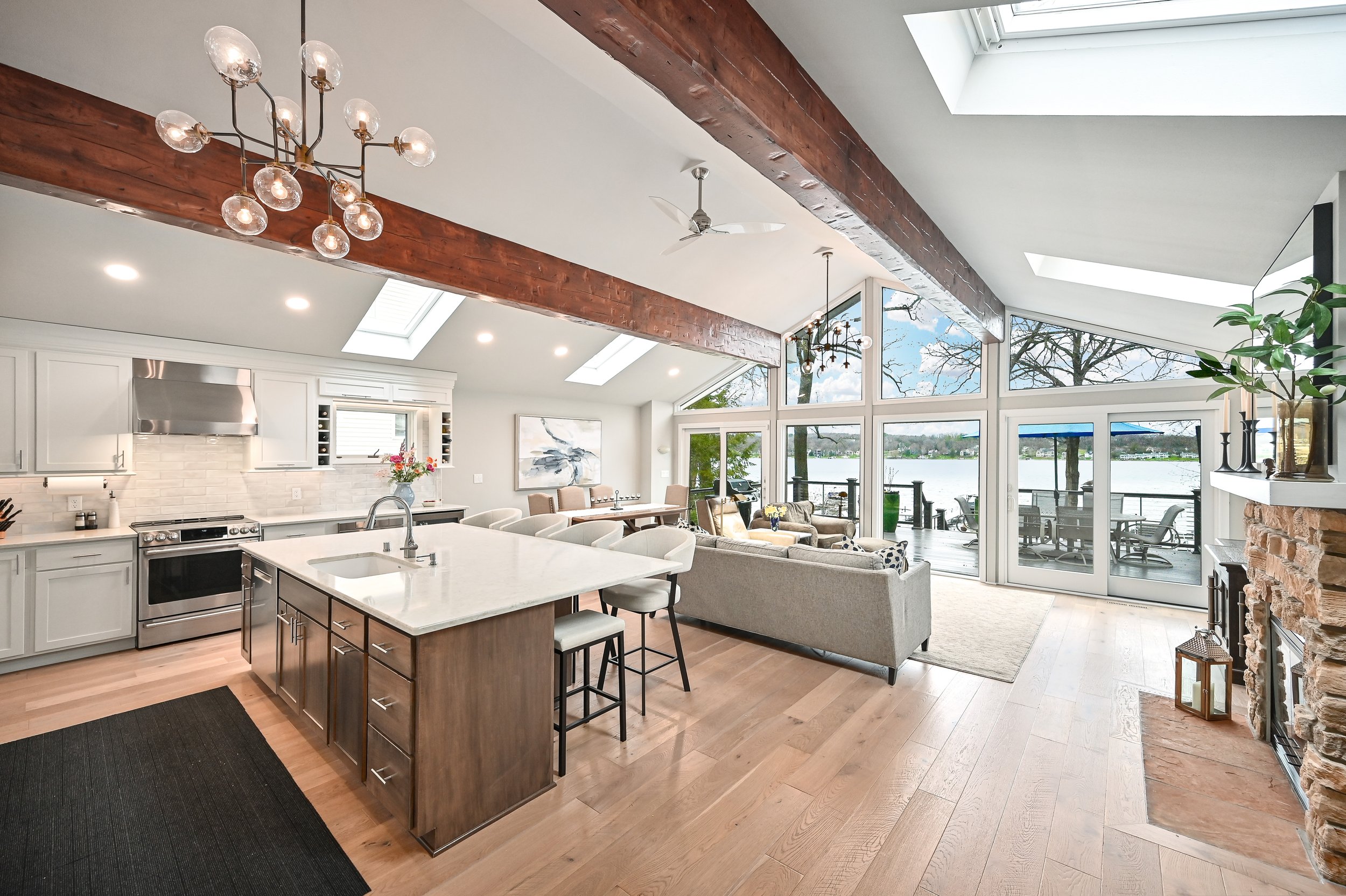 Spacious open-concept living area with kitchen, dining, and sitting spaces overlooking a lake through large windows, featuring hardwood floors, a stone fireplace, and modern furniture.