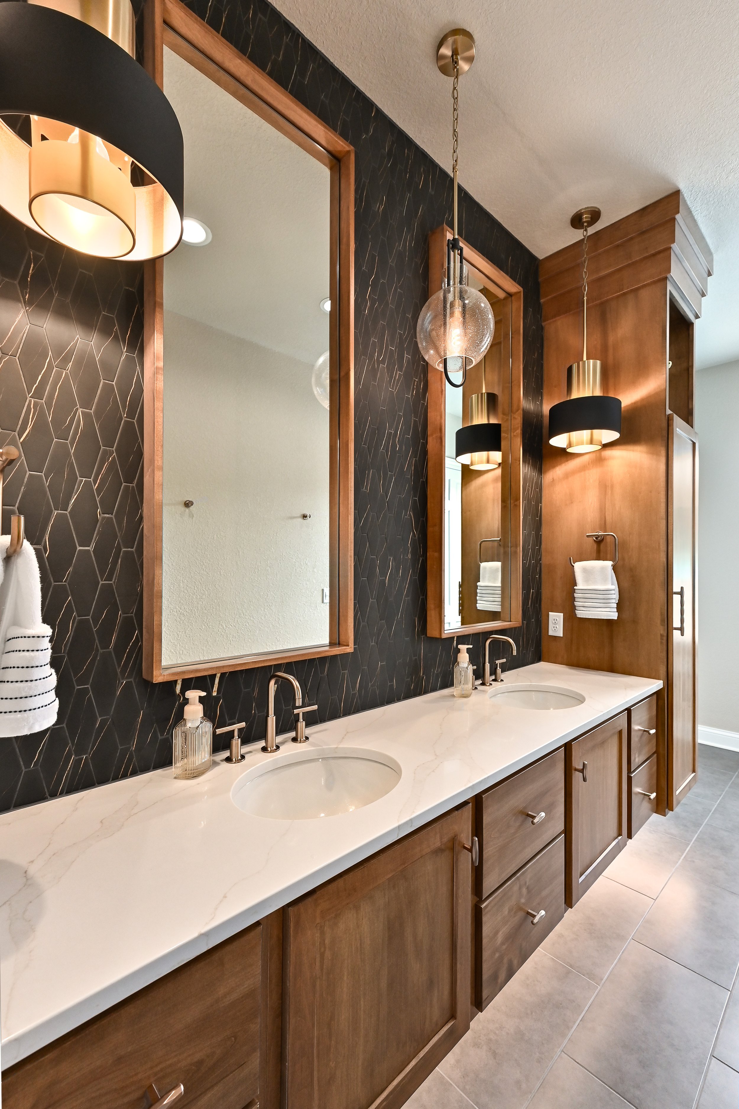 A double bathroom vanity with a white marble countertop, two oval undermount sinks, and wooden cabinets with silver handles. There are two large mirrors with wooden frames, black wall tiles with gold accents behind the sinks, and modern black and gol