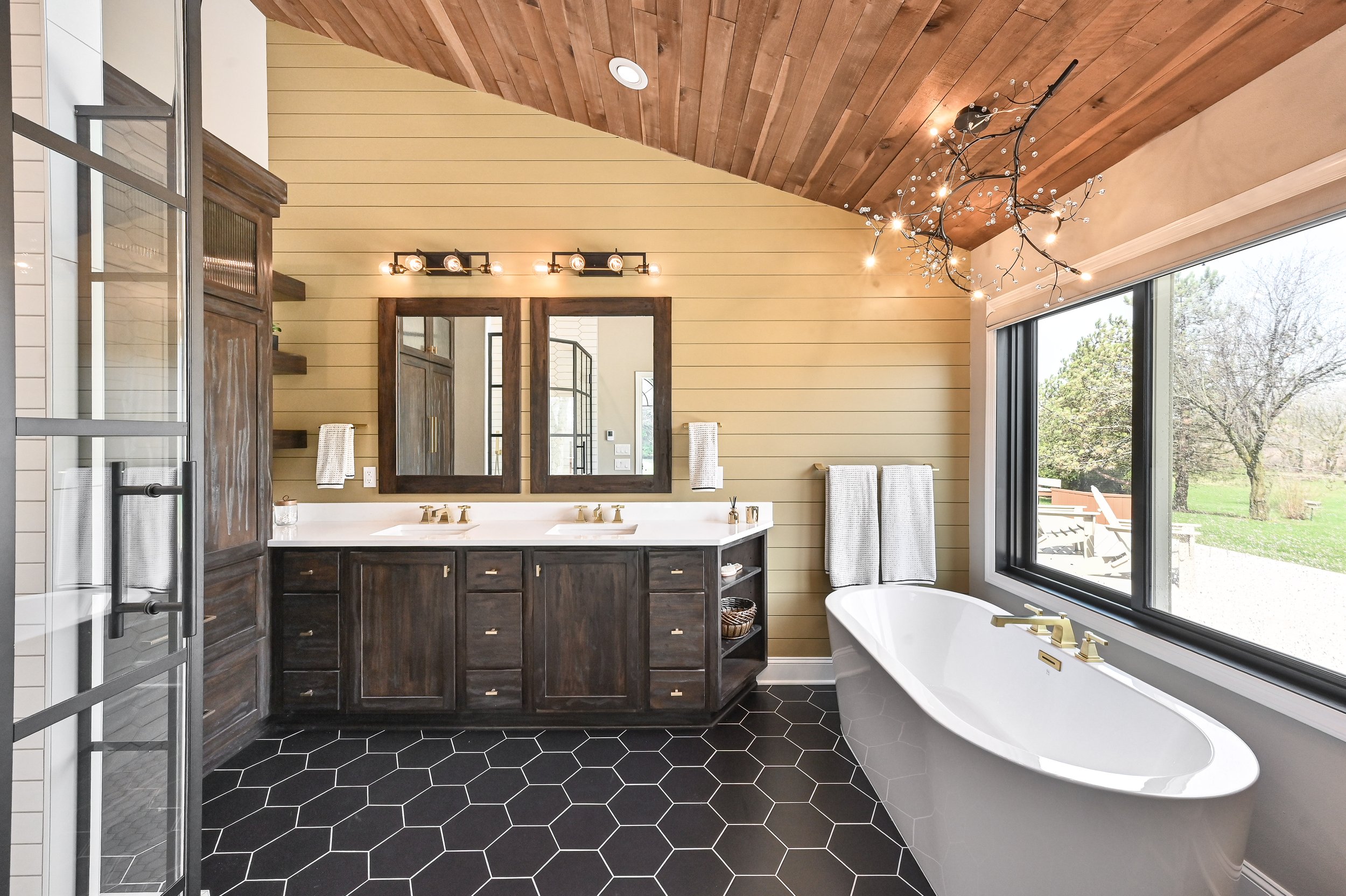 Modern bathroom with a double vanity, black hex tile floor, large bathtub near a big window, and wood-paneled ceiling with a decorative branch chandelier.