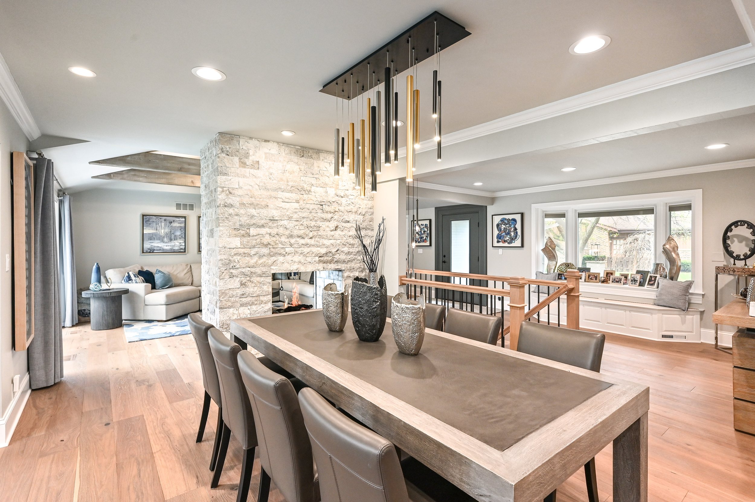 Open-concept living and dining area with a wooden dining table, modern pendant light, stone fireplace, comfortable seating, large window, and home decor.