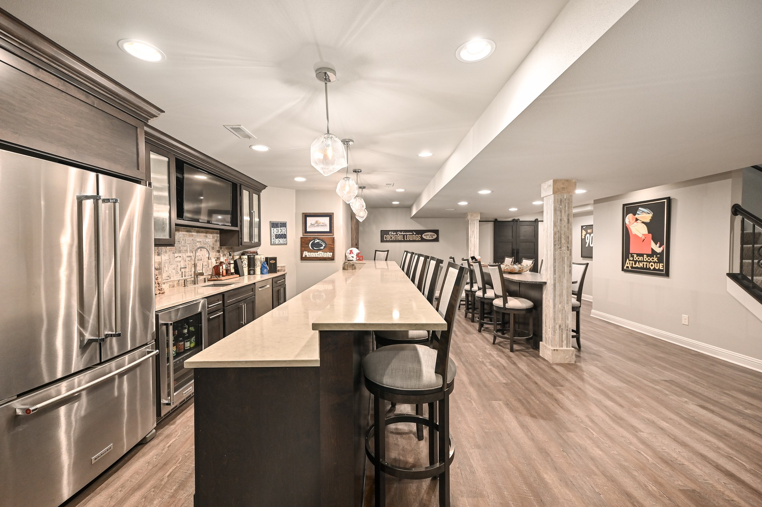 A modern basement bar with a long beige countertop, black cabinets, and stainless steel appliances. There are bar stools along the counter and framed posters on the walls.