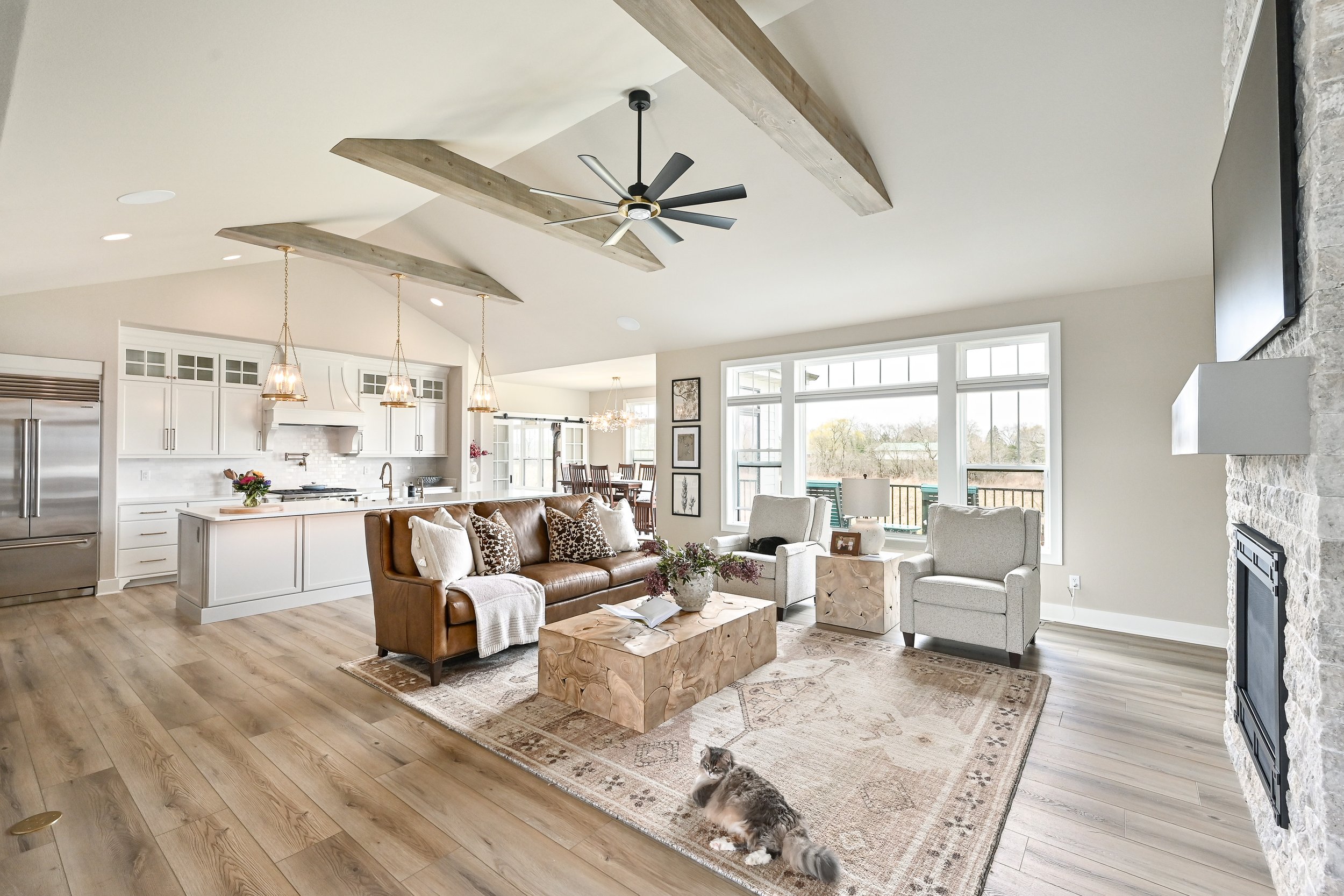 Open-concept living room and kitchen with wood flooring, white cabinets, a brown leather sofa, white armchairs, a stone fireplace, and large windows showing outdoor views.
