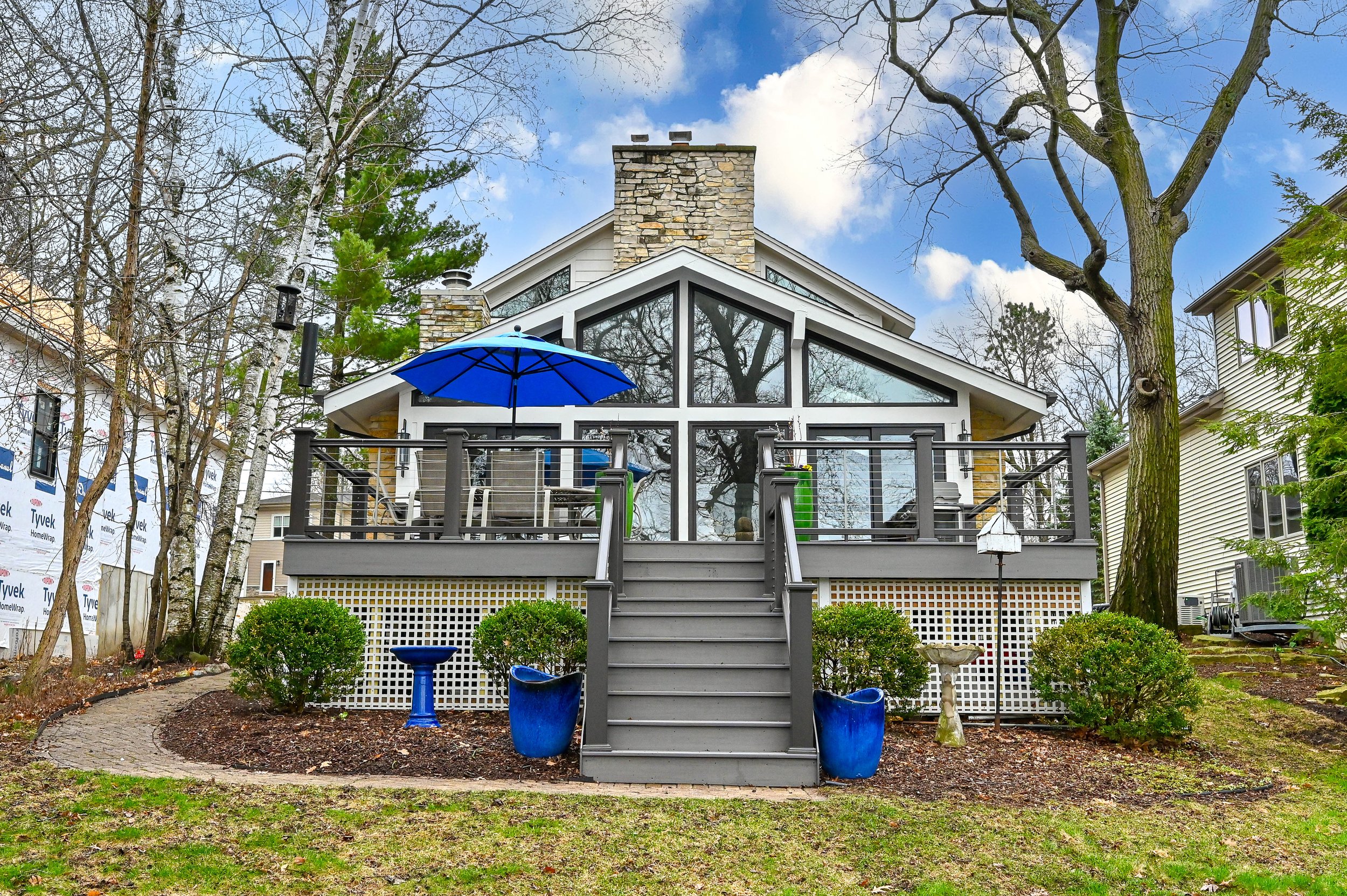 A modern house with a large glass front and a wooden deck with an umbrella, surrounded by trees and bushes, and a stone chimney.