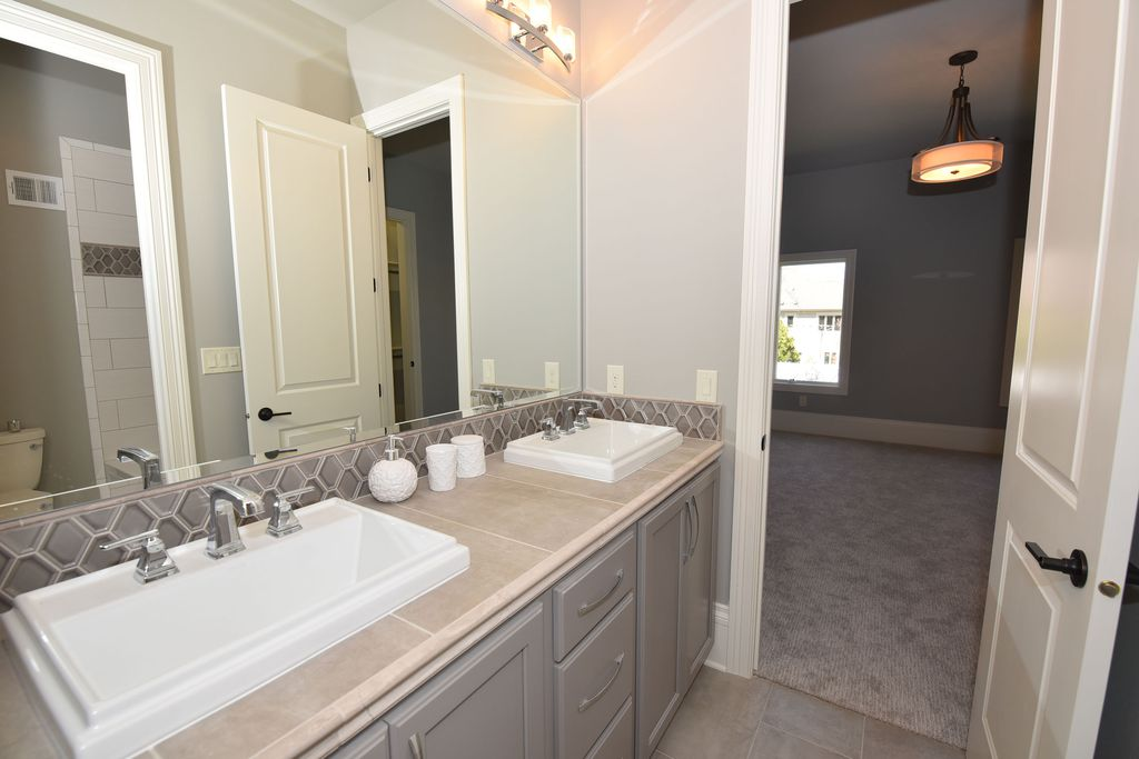A bathroom vanity with two sinks, a large mirror, and decorations such as white vases and containers, with a doorway leading to a bedroom and a view of a room with a window and ceiling light.