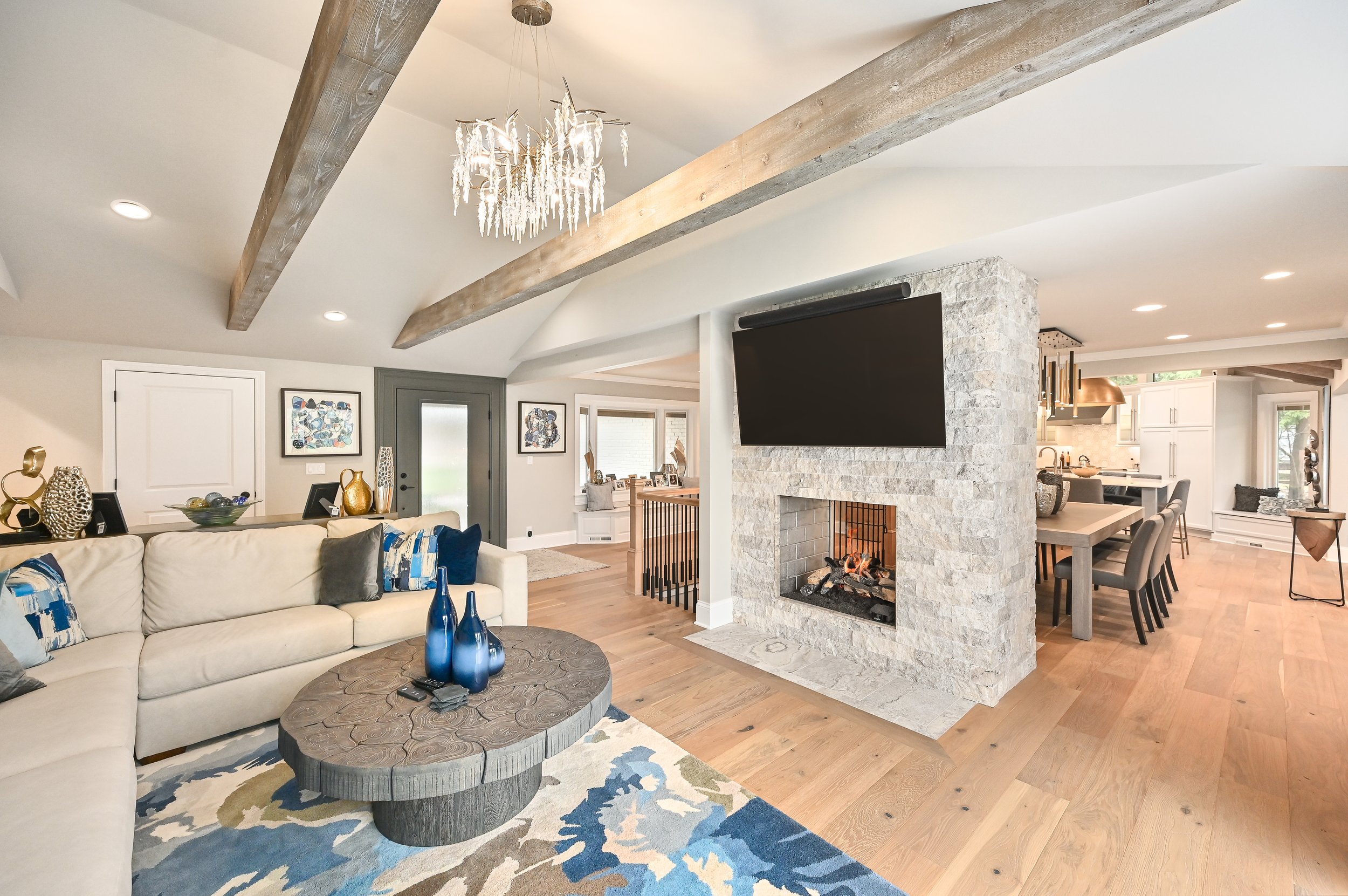 Open-concept living room with a beige sectional sofa, a wooden coffee table with blue vases, a stone fireplace with a mounted flat-screen TV, hardwood floors, and a chandelier, leading to a dining area and kitchen.