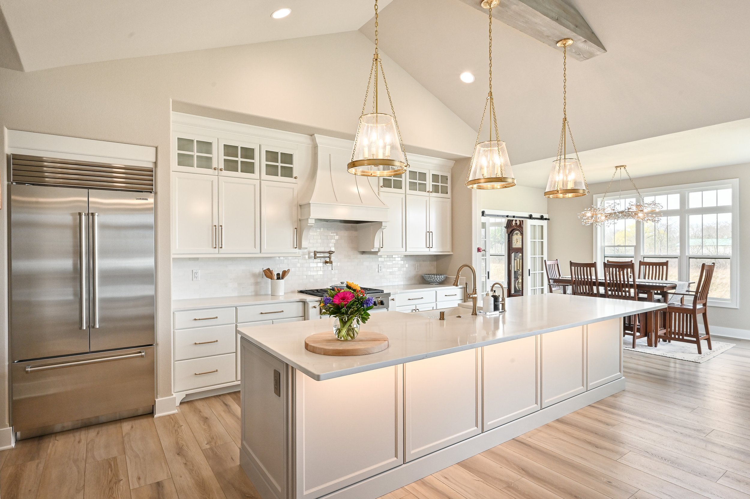 Modern kitchen with white cabinets, a large island with a white countertop, stainless steel refrigerator, and a dining area with wooden chairs and large windows.