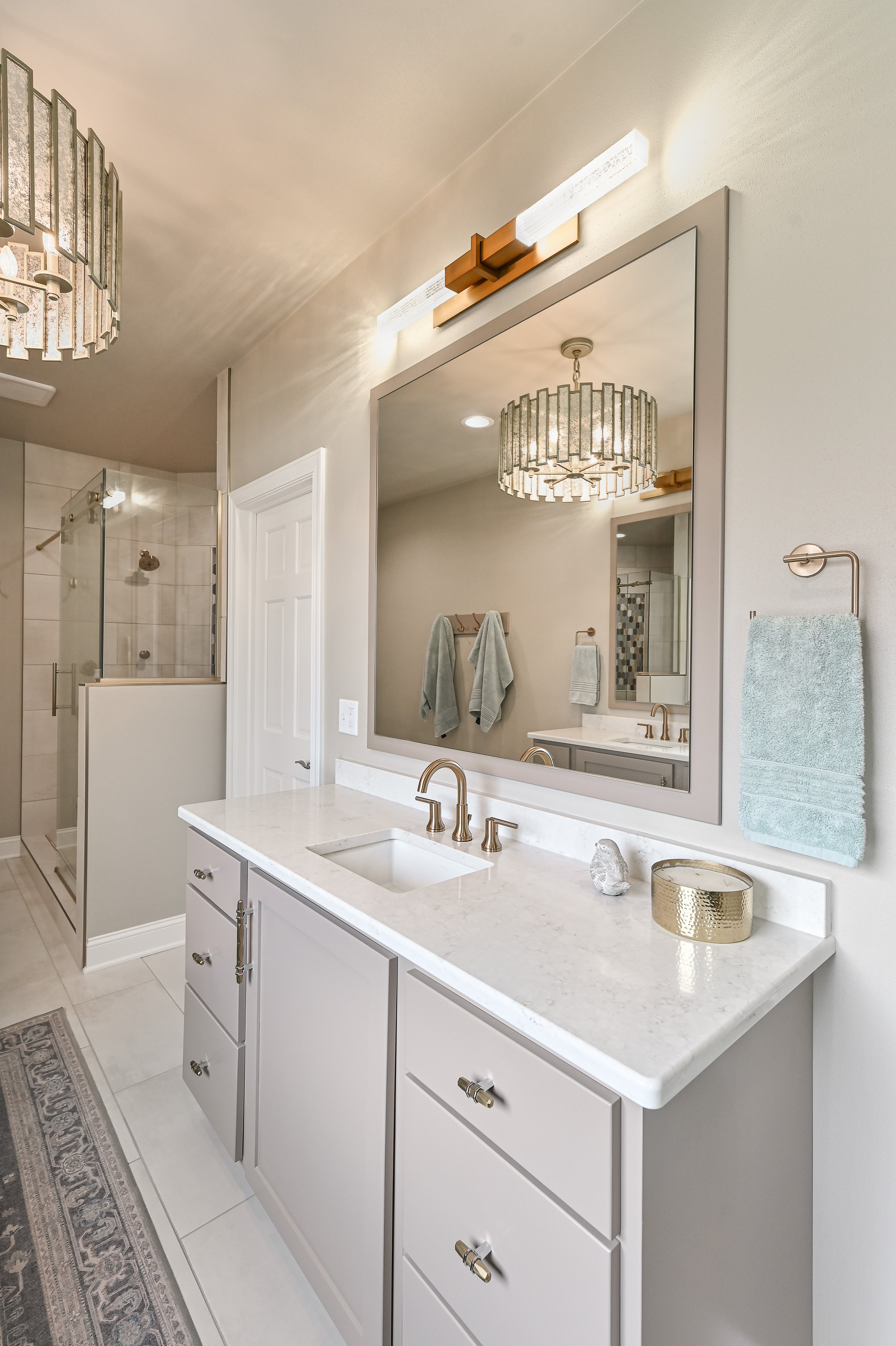 Modern bathroom vanity with white cabinets, a marble countertop, and a gold faucet. Large mirror above the sink, decorated with a small vase and a round gold container. Towel rack with a folded towel. Seen through a reflection, there is a separate sh