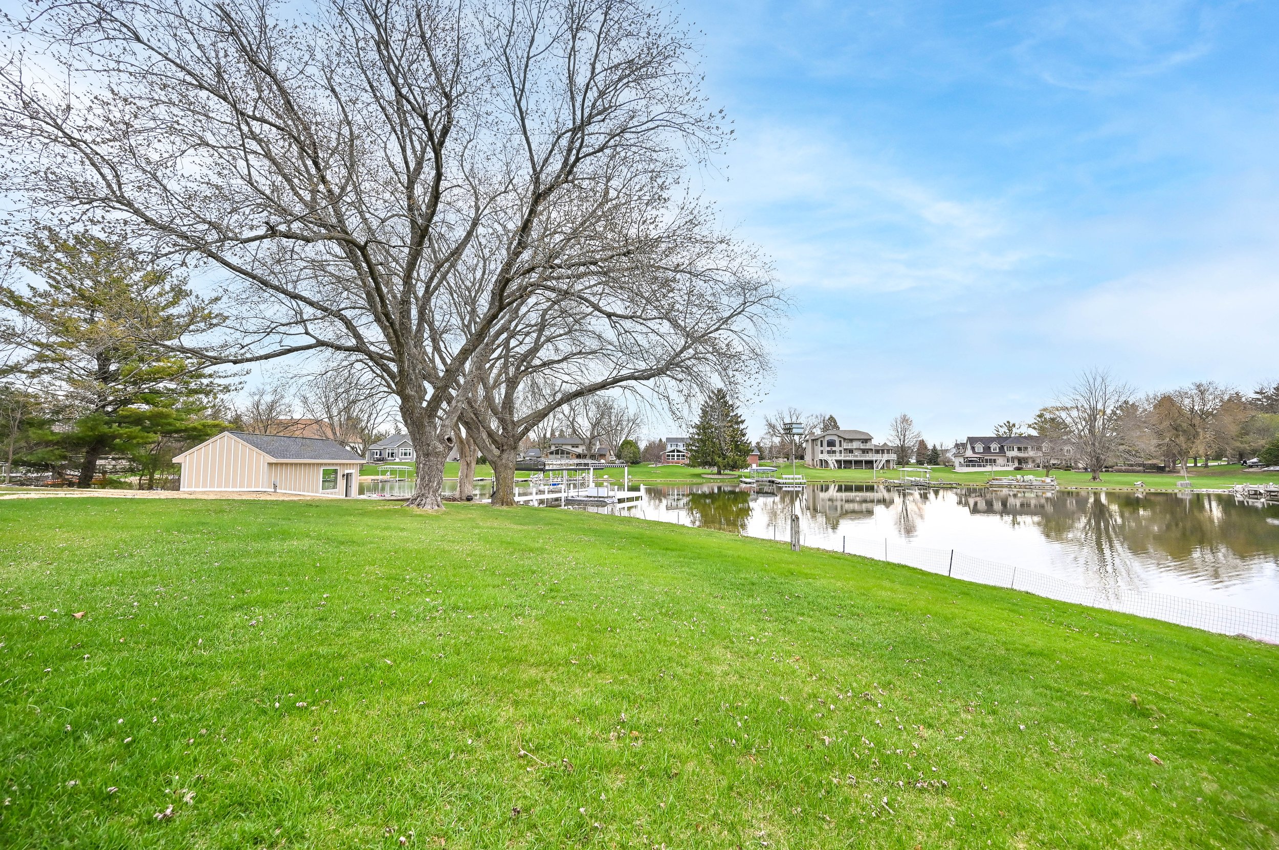 A scenic view of a suburban neighborhood by a calm river with green grass, large leafless trees, and houses in the background under a partly cloudy sky.
