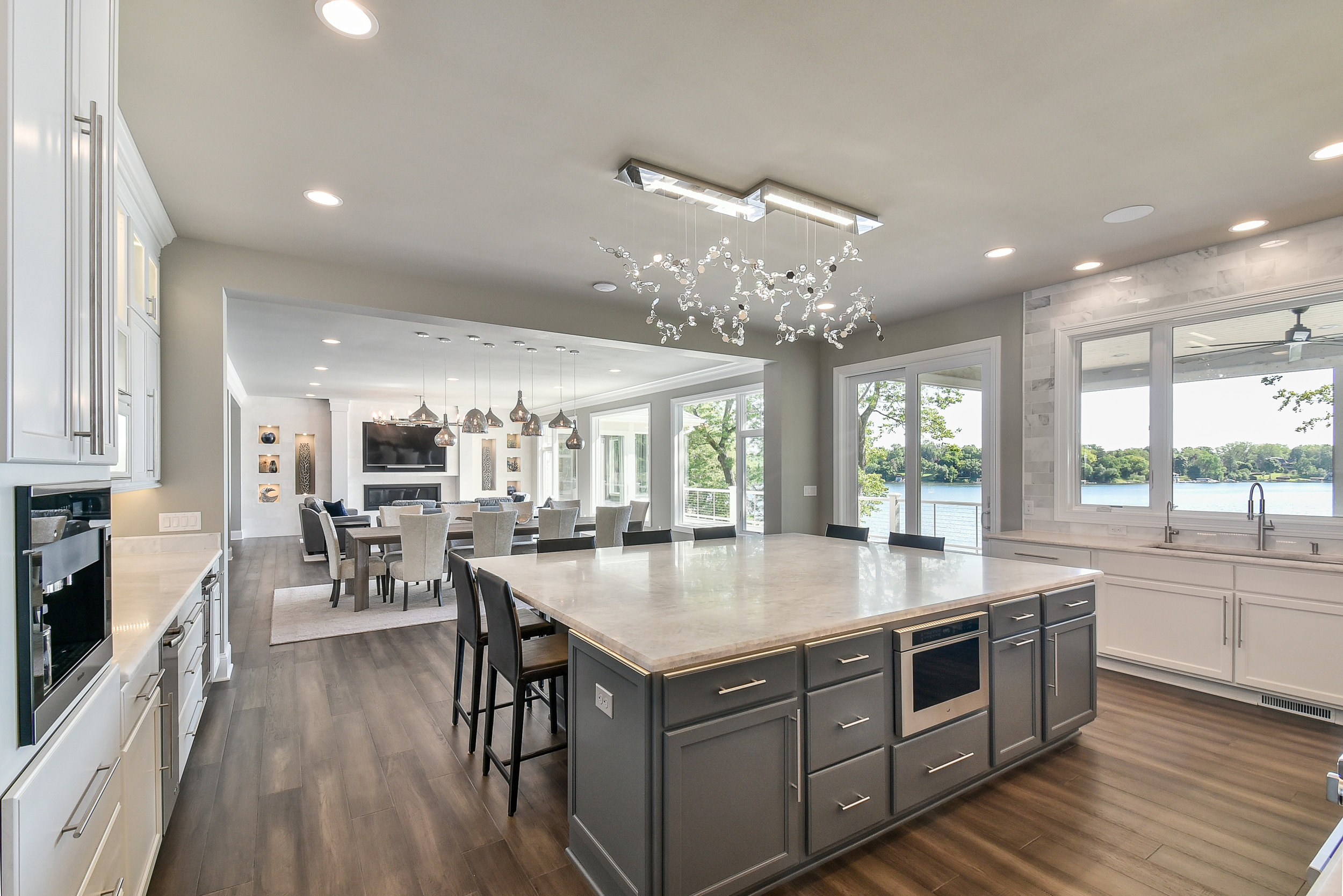 Open-concept kitchen and dining area with large island, modern lighting fixtures, and a view outside over a lake.