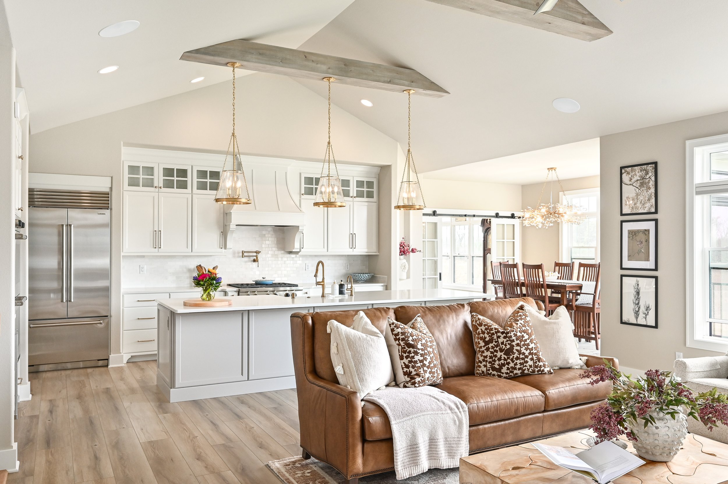 Open-plan living room and kitchen with white cabinetry, wooden flooring, brown leather sofa decorated with pillows, kitchen island with a vase of flowers, dining area with wooden table and chairs, large windows, and contemporary lighting fixtures.