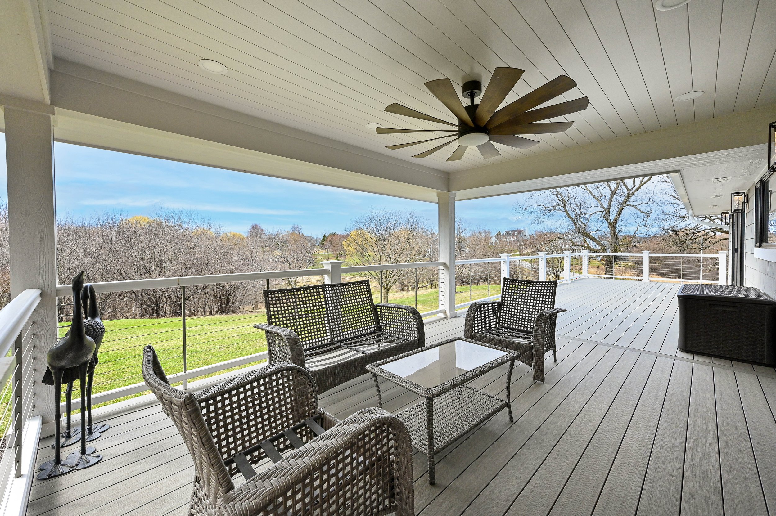 Covered outdoor deck with wicker furniture, a coffee table, a ceiling fan, and a landscape view of trees and a lawn