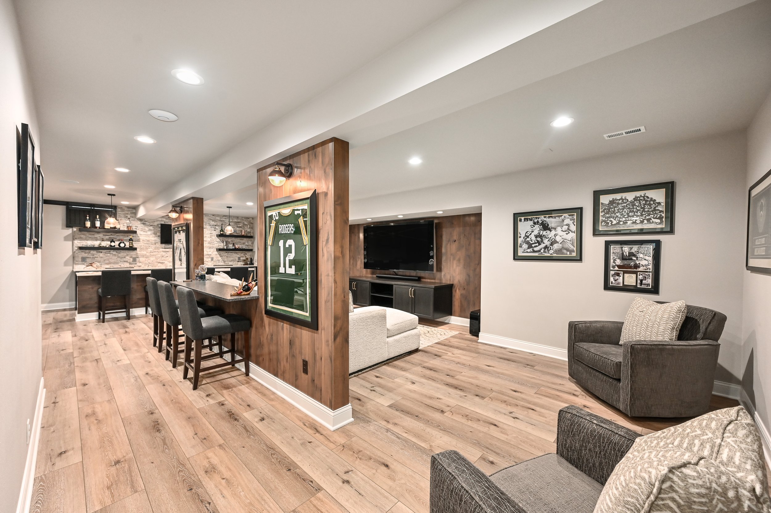 View of a cozy basement lounge with a large flat-screen TV, framed sports memorabilia, a sitting area with chairs and a couch, a wooden bar counter with stools, and a stone accent wall in the background.