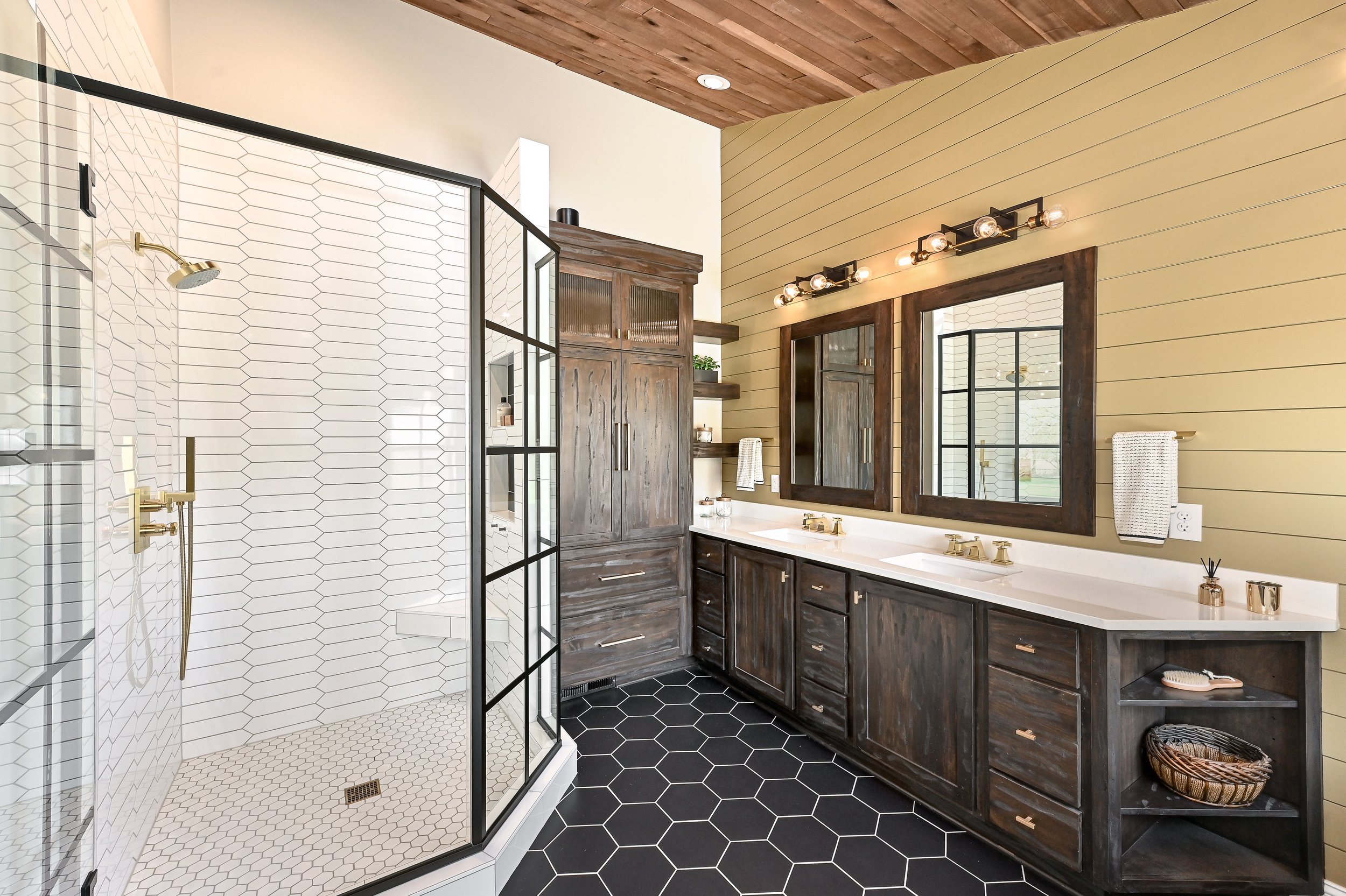 Modern bathroom with a walk-in shower featuring white and black hexagon tiles, a double vanity with dark wood cabinets, and a wooden framed mirror. The room has a wood-paneled ceiling and a yellow shiplap wall.