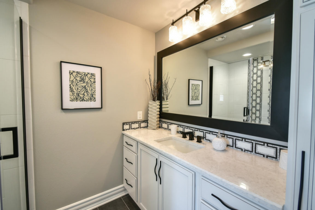 Bathroom vanity with a large mirror, decorative black and white tile border, and a vase with dried branches.