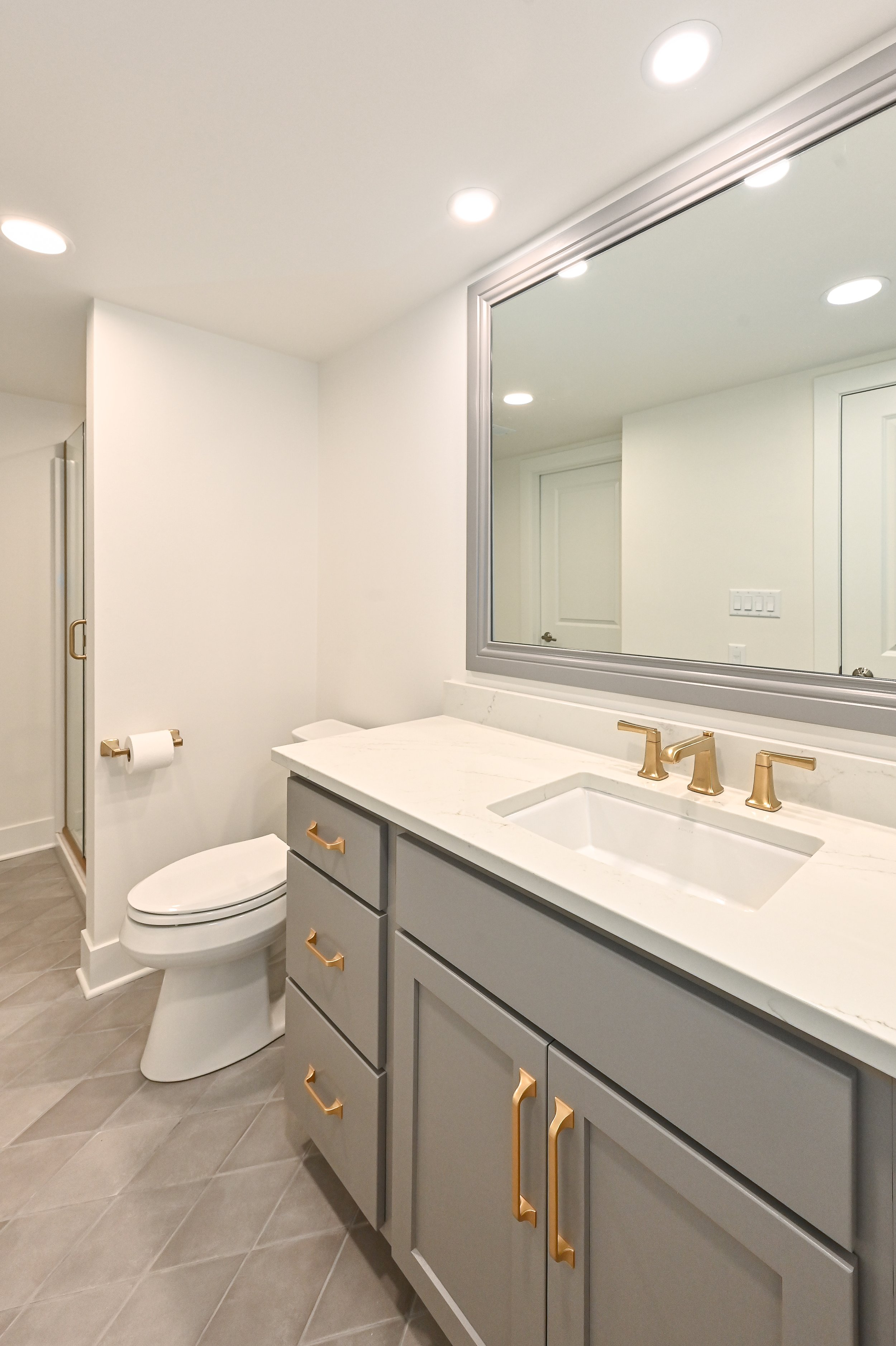 Modern bathroom with white walls, gray cabinetry with gold handles, a large mirror, a white marble countertop with a built-in sink, gold fixtures, and a toilet. There is a shower with a glass door in the corner.