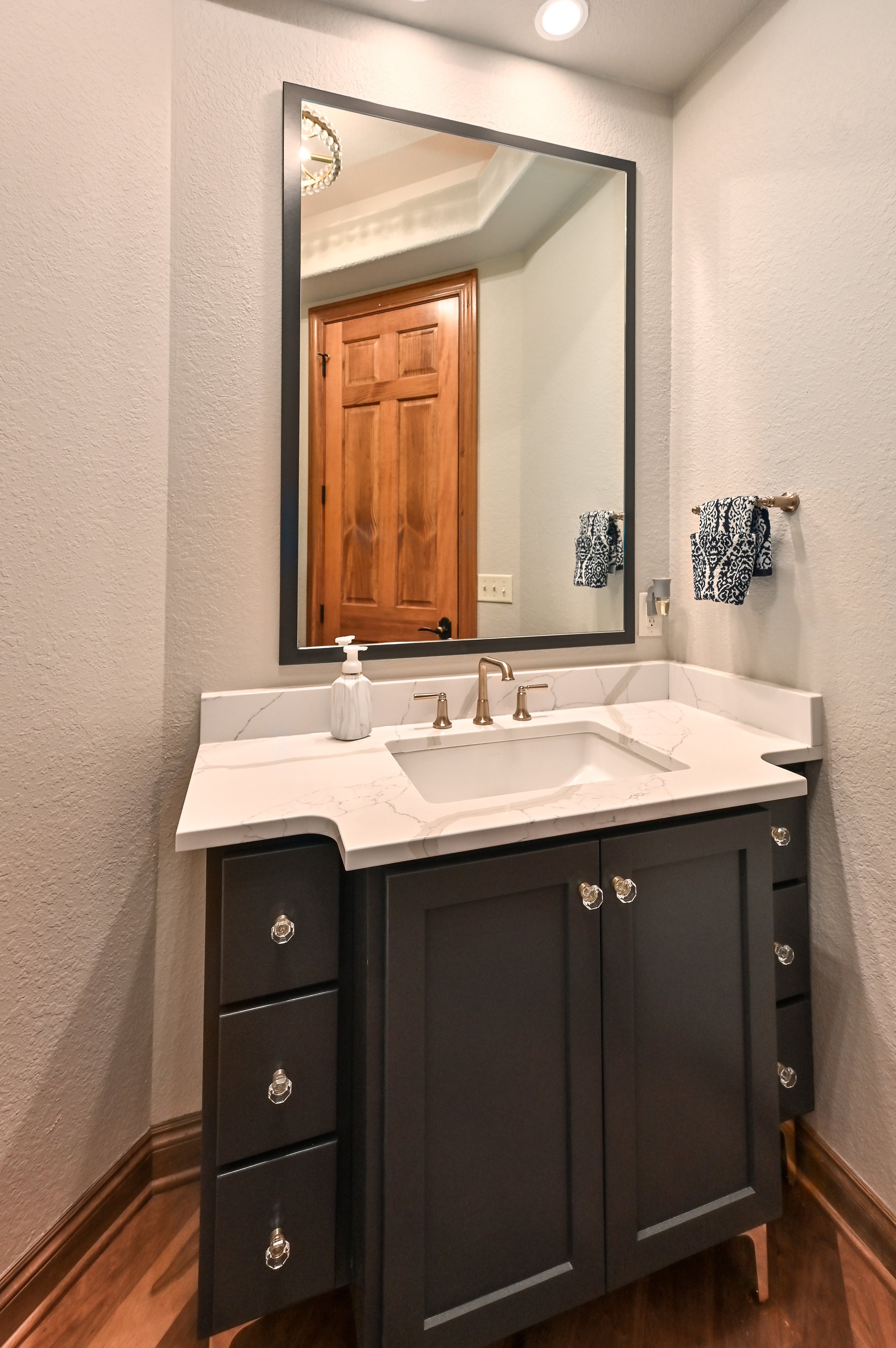 Bathroom vanity with a dark cabinet, white marble countertop, and a large mirror. Towel rack with two blue and white patterned towels. Wooden door reflected in the mirror.