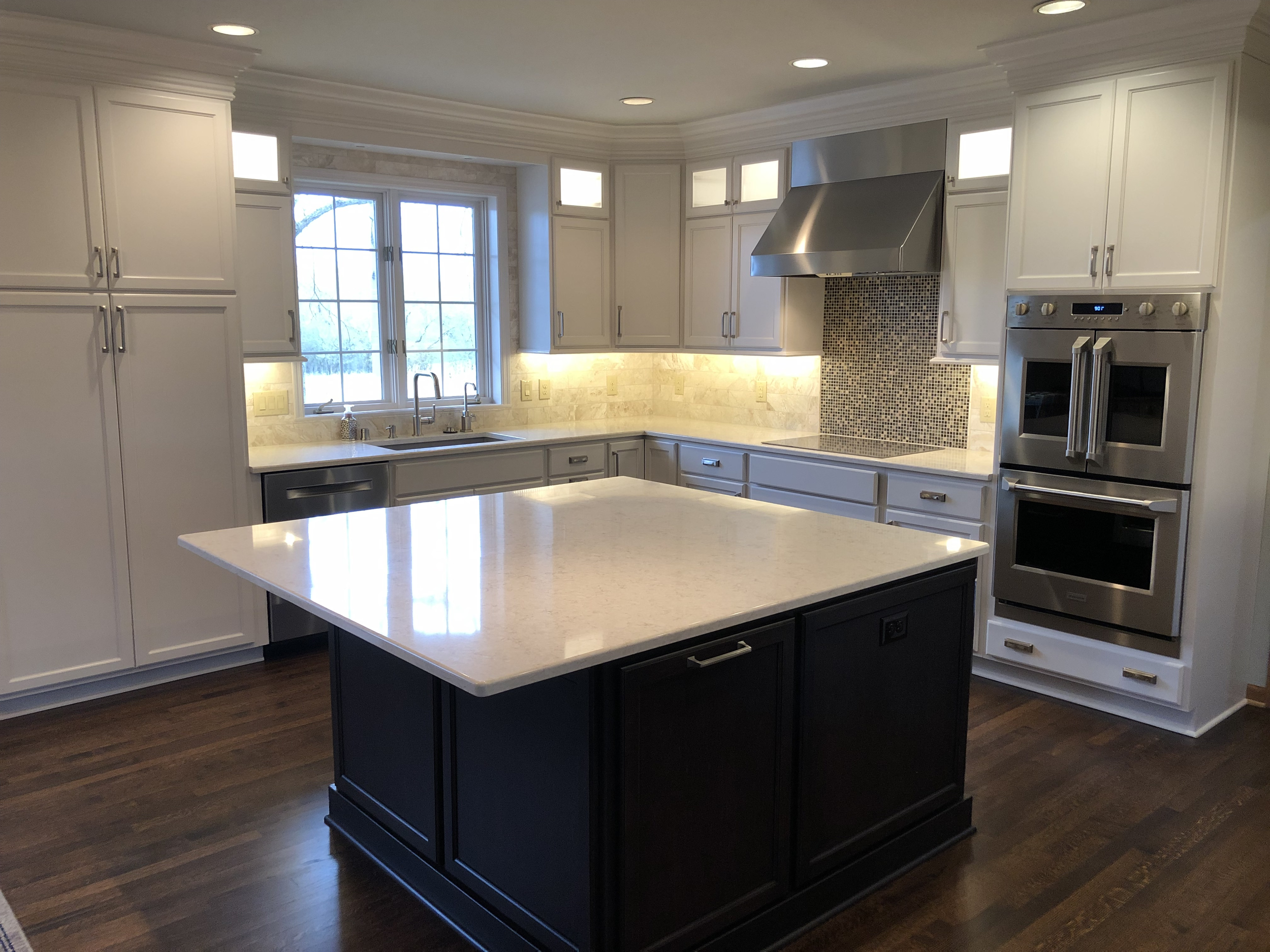 Modern kitchen with white cabinets, black island, marble countertops, stainless steel oven and microwave, range hood, window above sink, dark wood flooring.