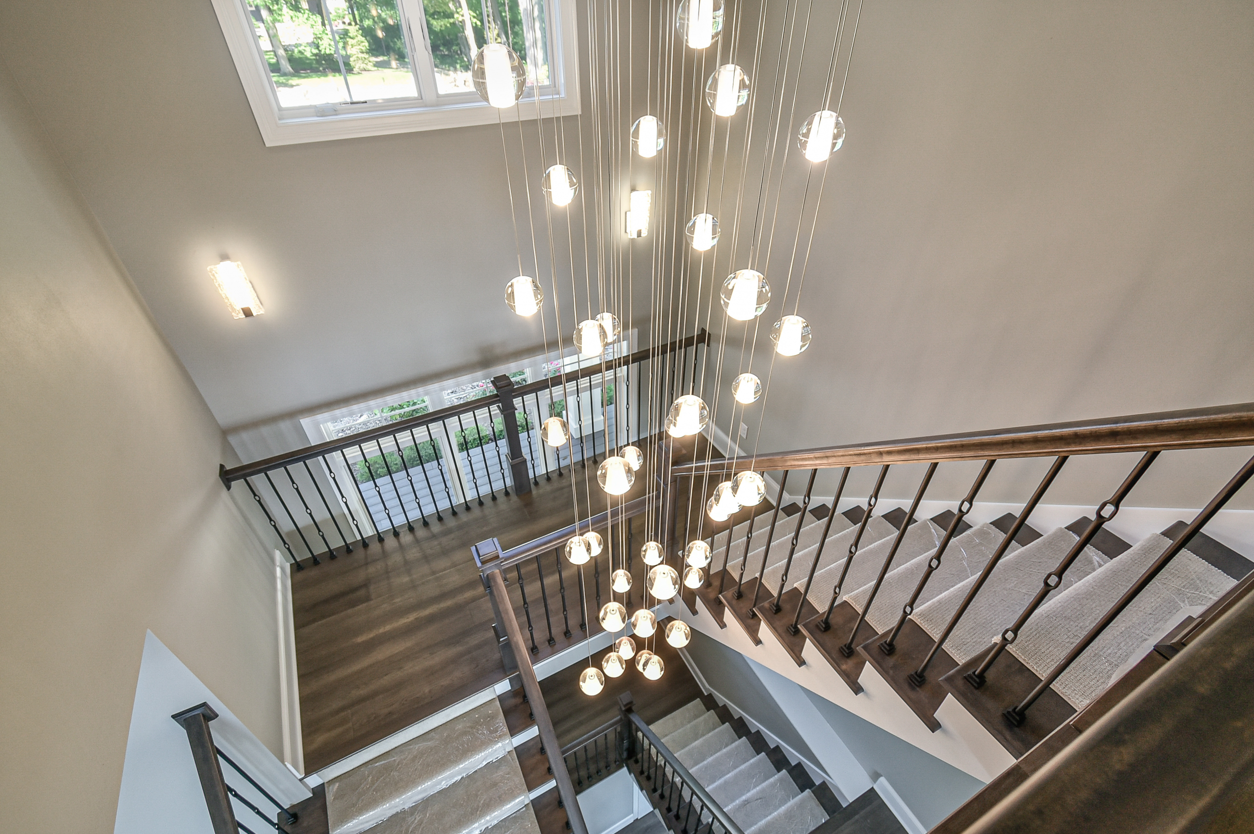 Interior view of a staircase with a modern chandelier, wooden stairs, black iron railings, and large windows allowing natural light.