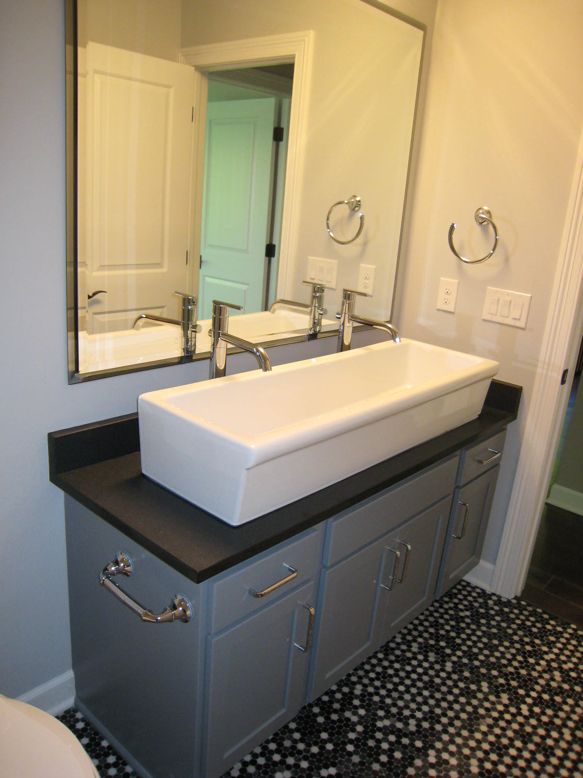 Bathroom vanity with a rectangular vessel sink, chrome faucets, large wall mirror, gray cabinet, and black and white mosaic tile floor.